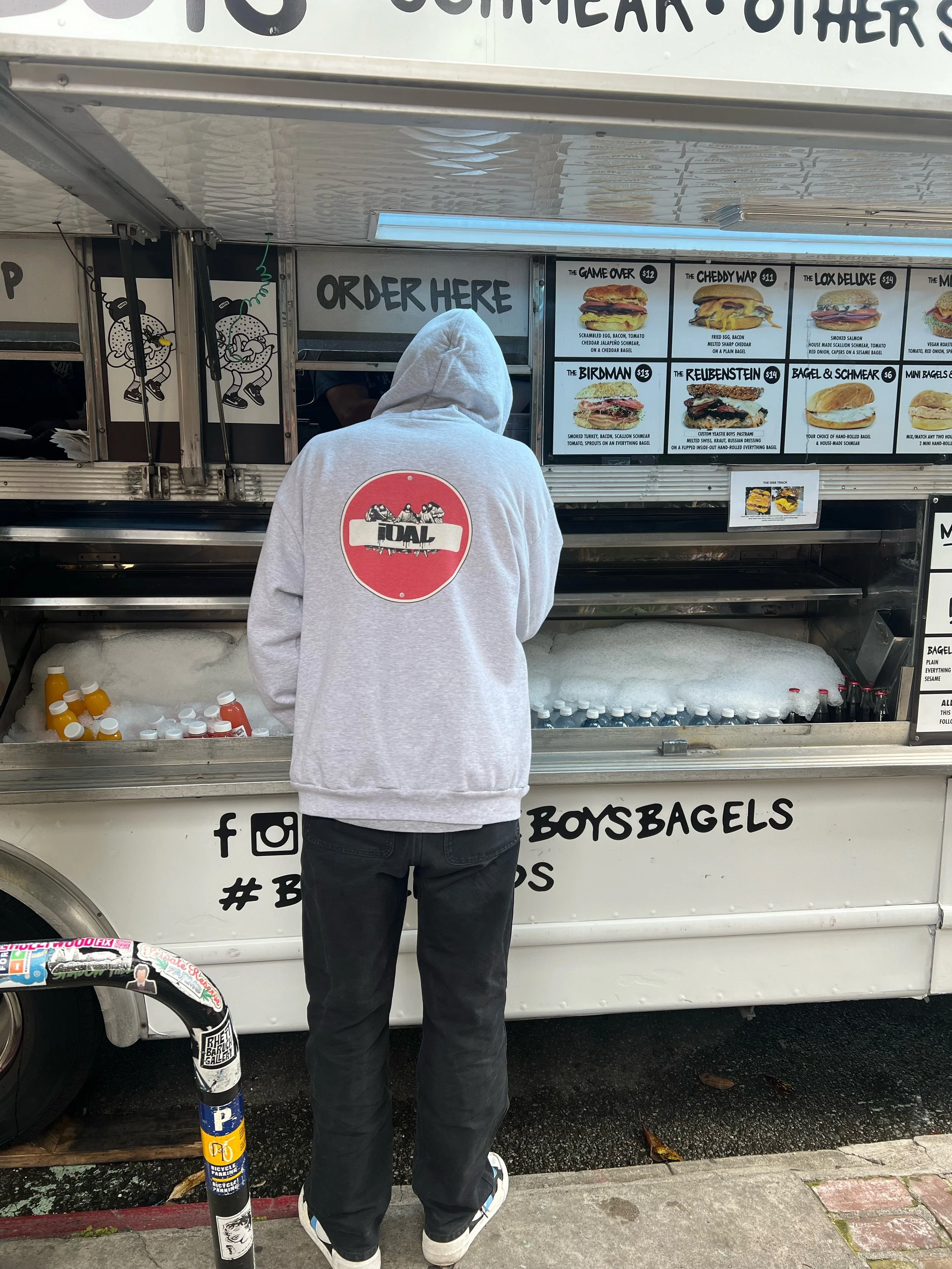 A person in a grey hoodie stands at a food truck counter, looking at a menu displaying various burger options.