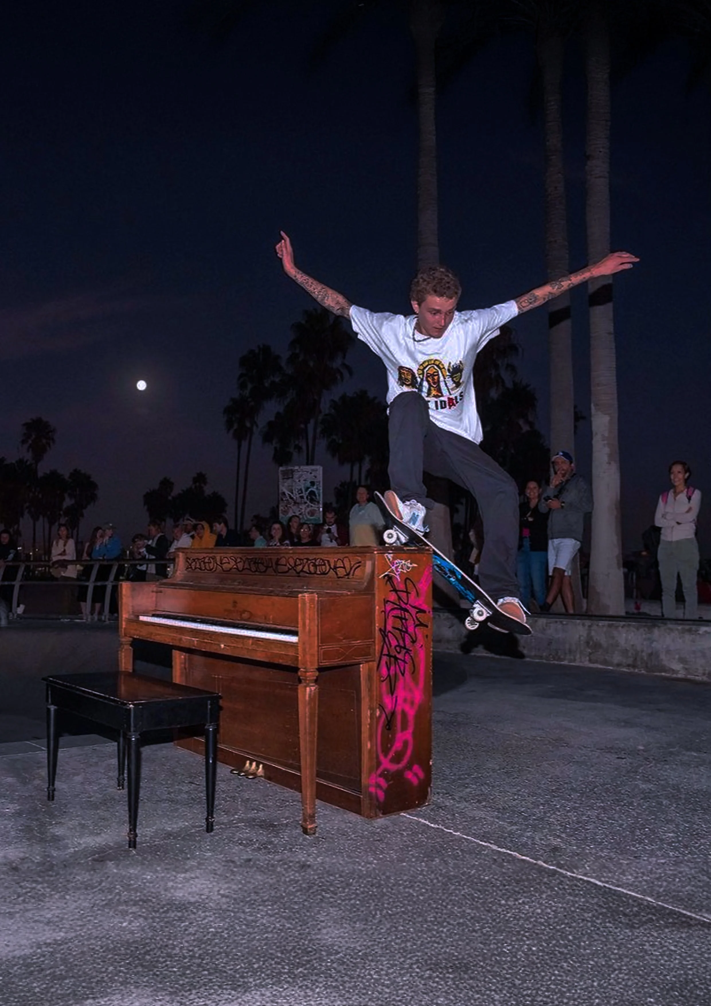 Skateboarder performing a trick over an old piano at night with palm trees and a full moon in the background. Spectators watch from behind.