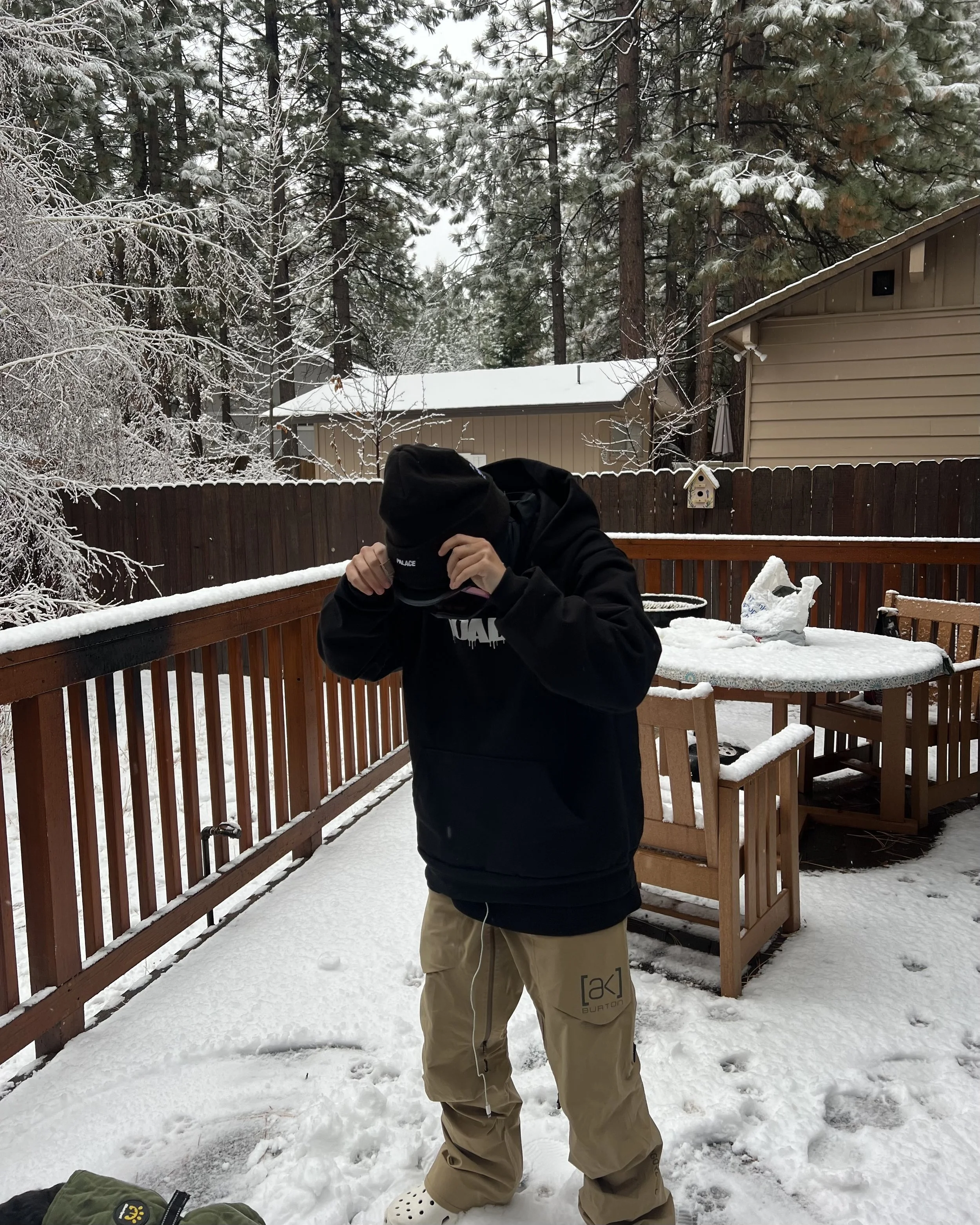 A person in a black hoodie, beige pants, and a black cap standing outside on a snowy deck, with trees and houses in the background.