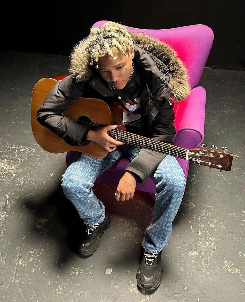 Young man with blonde dreadlocks sitting on a purple chair, holding an acoustic guitar, wearing a black jacket with fur trim and jeans with a repeating pattern, in a dimly lit room.