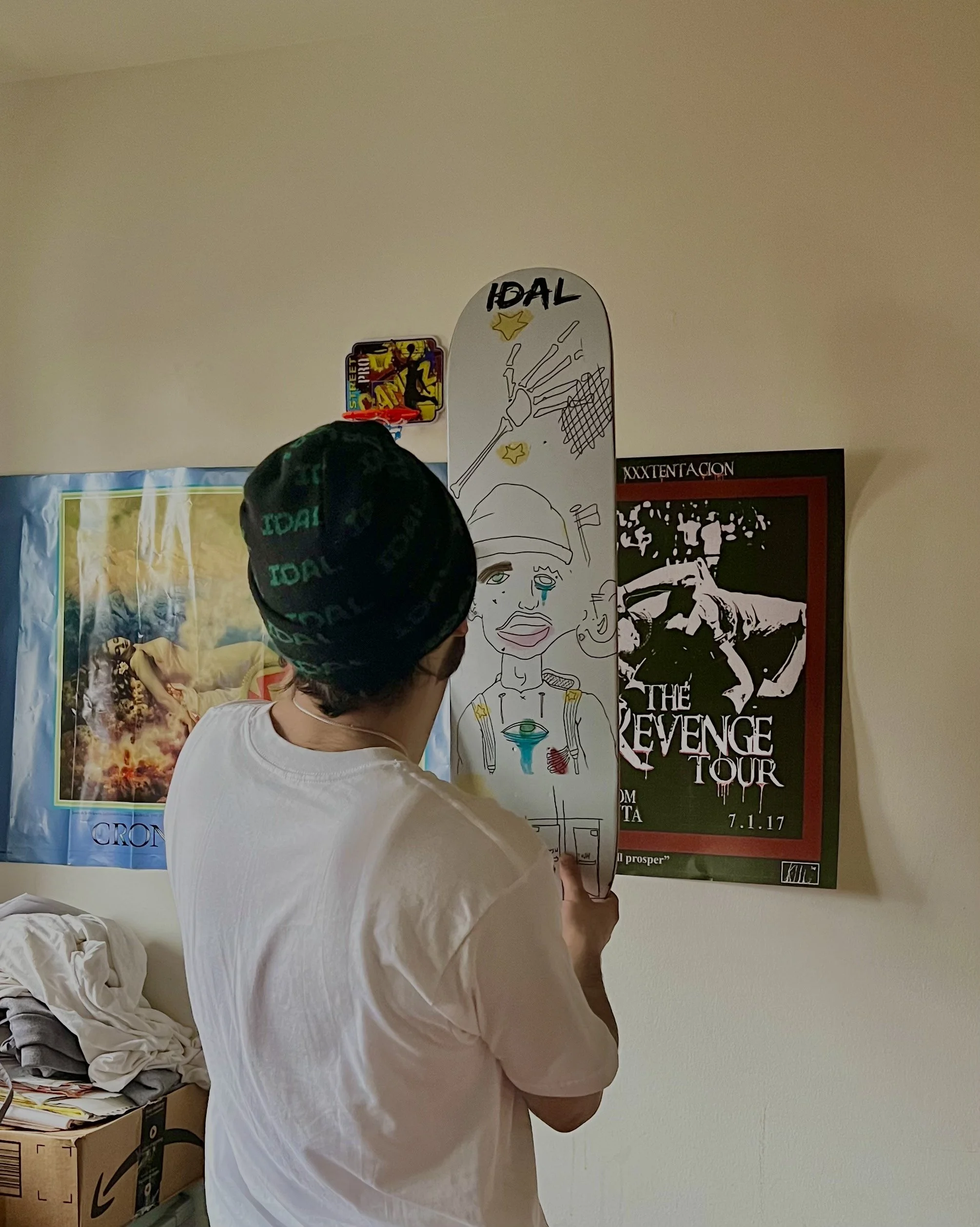A person wearing a black beanie and a white t-shirt is drawing on a skateboard-shaped board that has the word 'IDAL' and a cartoon face with tears and various doodles. The person is standing in a room with posters on the wall, including one with a bl