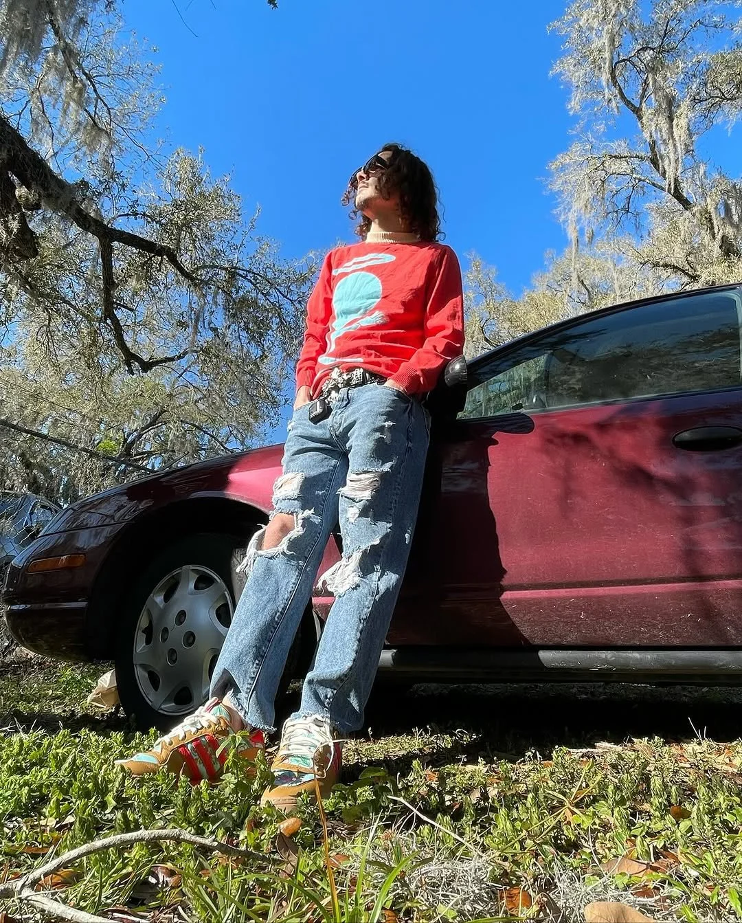 Person leaning against a red car outdoors on a sunny day, wearing a red sweater with a blue and white graphic, distressed jeans, and colorful sneakers, with trees and a bright blue sky in the background.
