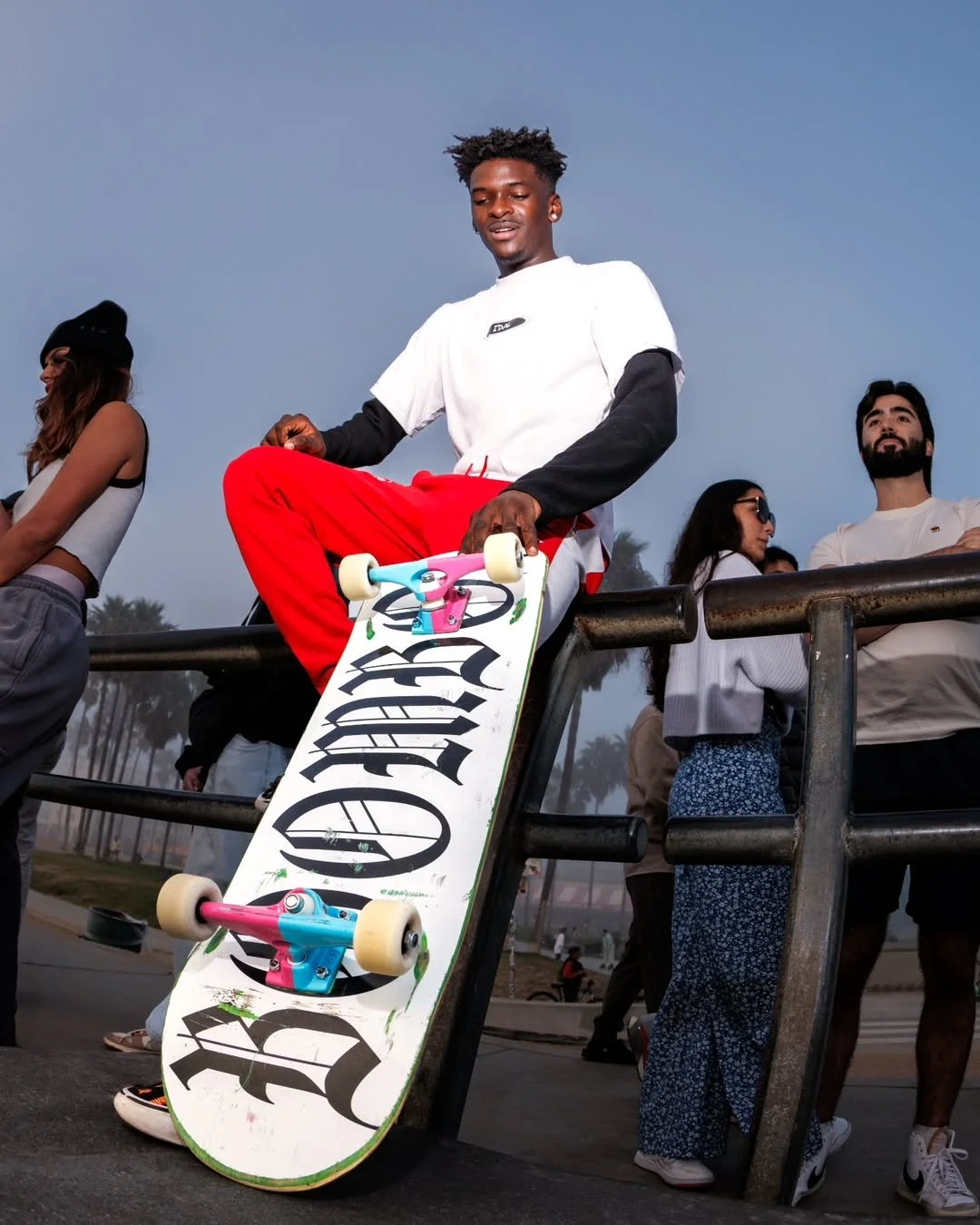A young man sitting on a railing with a skateboard hanging off his lap, outdoors during dusk. There are several people in the background.