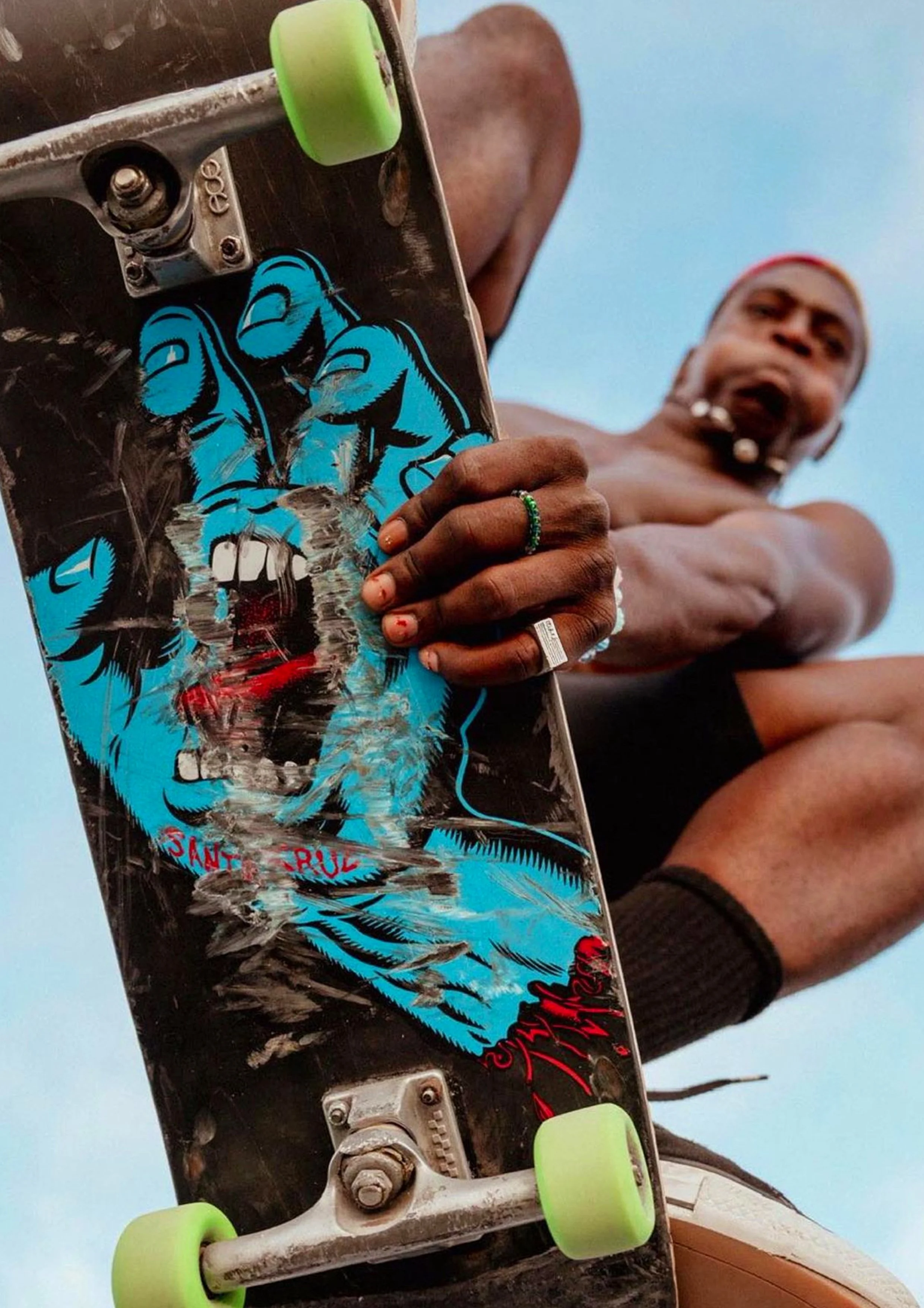 A shirtless man sitting cross-legged outdoors holding a skateboard with a blue and black graphic of a snarling animal, flames, and text. The man wears rings and beaded jewelry, and a beanie hat, with a blue sky in the background.