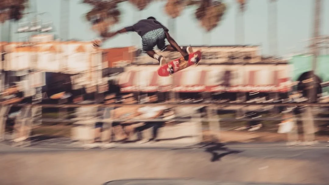 Skateboarder performing an aerial trick at a skate park with palm trees and spectators in the background.