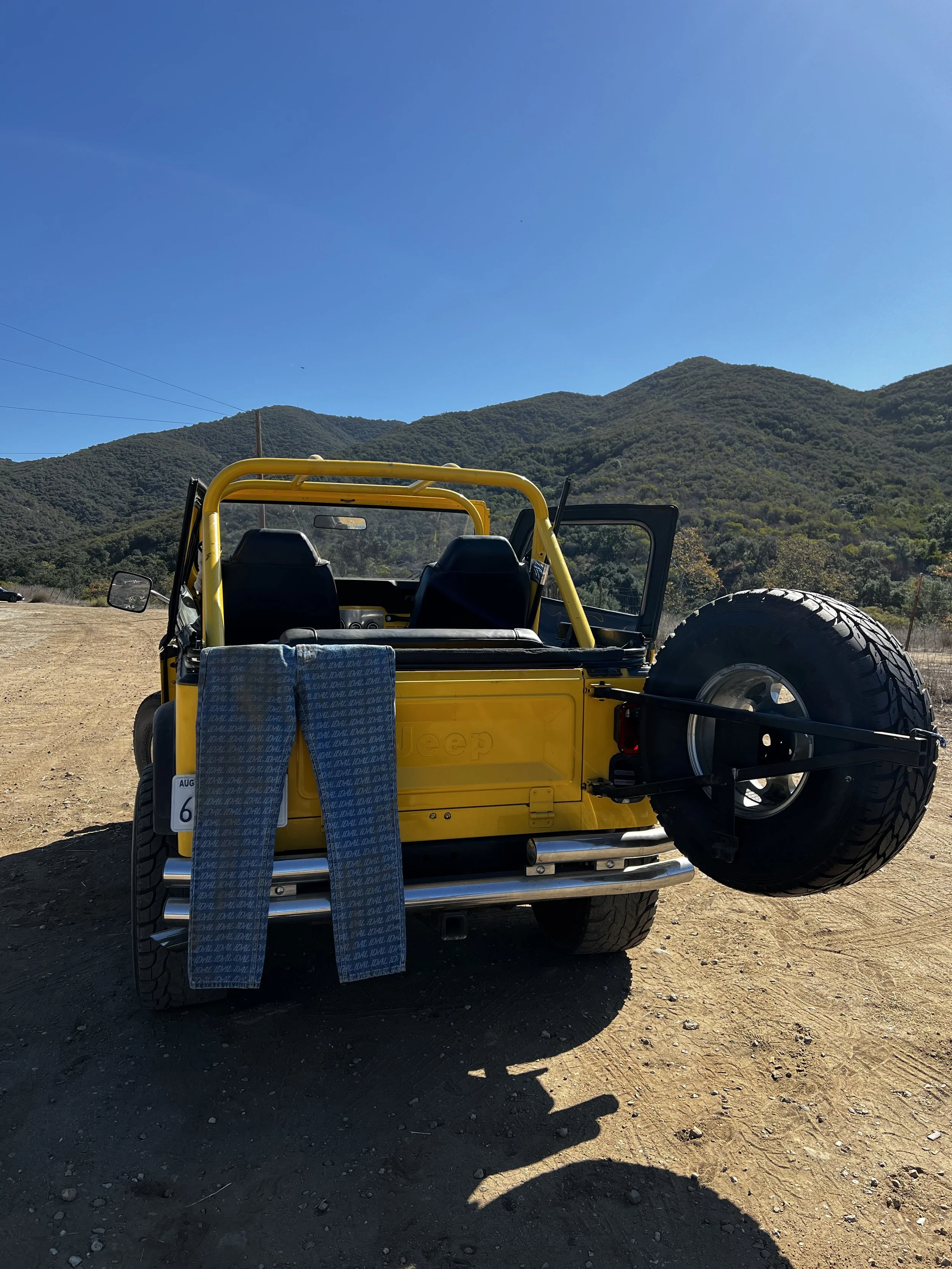 Yellow Jeep parked on a dirt lot with mountains in the background, a spare tire mounted on the rear, and two towels hanging from the back.
