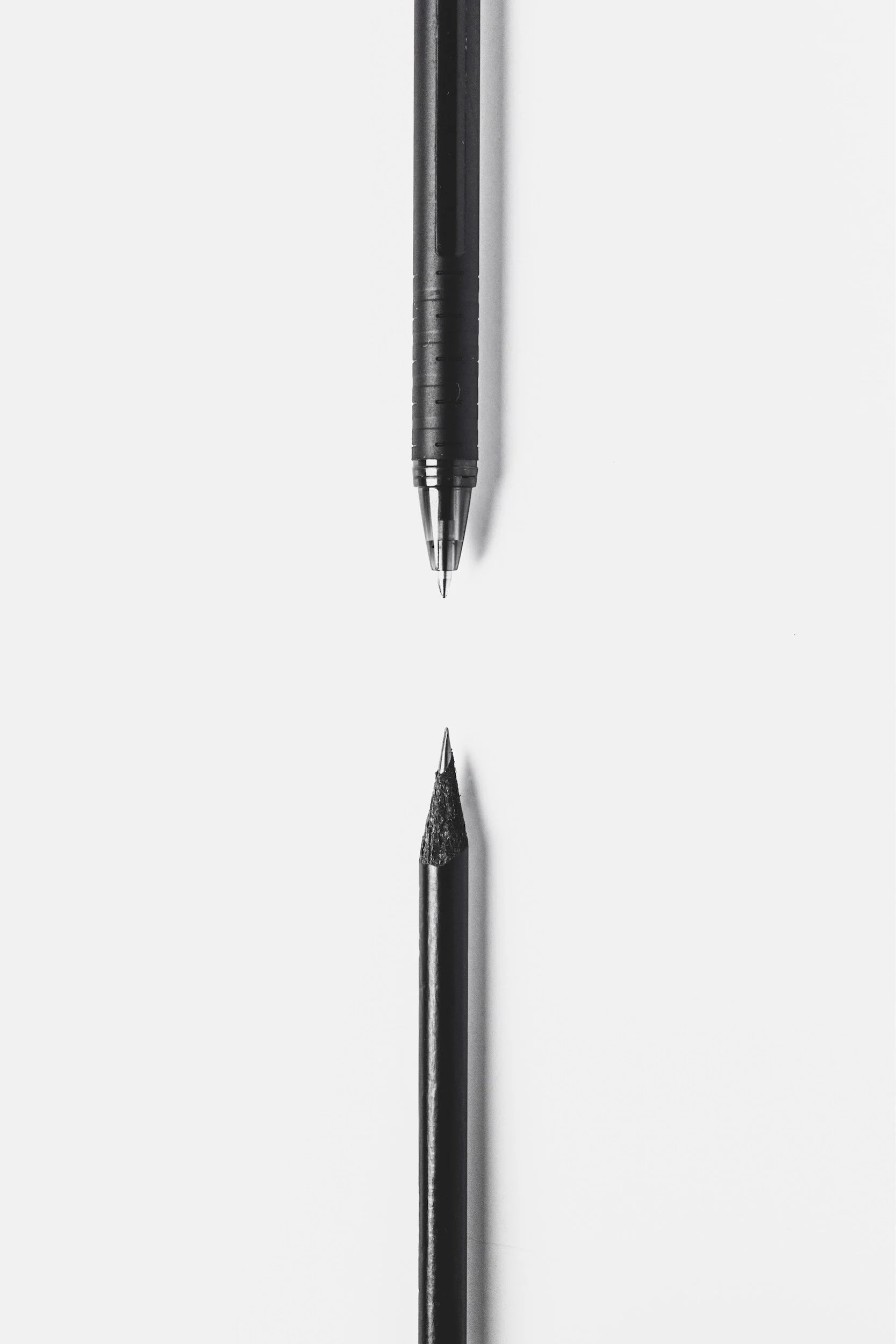 A black ballpoint pen facing downward and a black pencil facing upward are positioned close to each other on a white background. The pen is above the pencil.