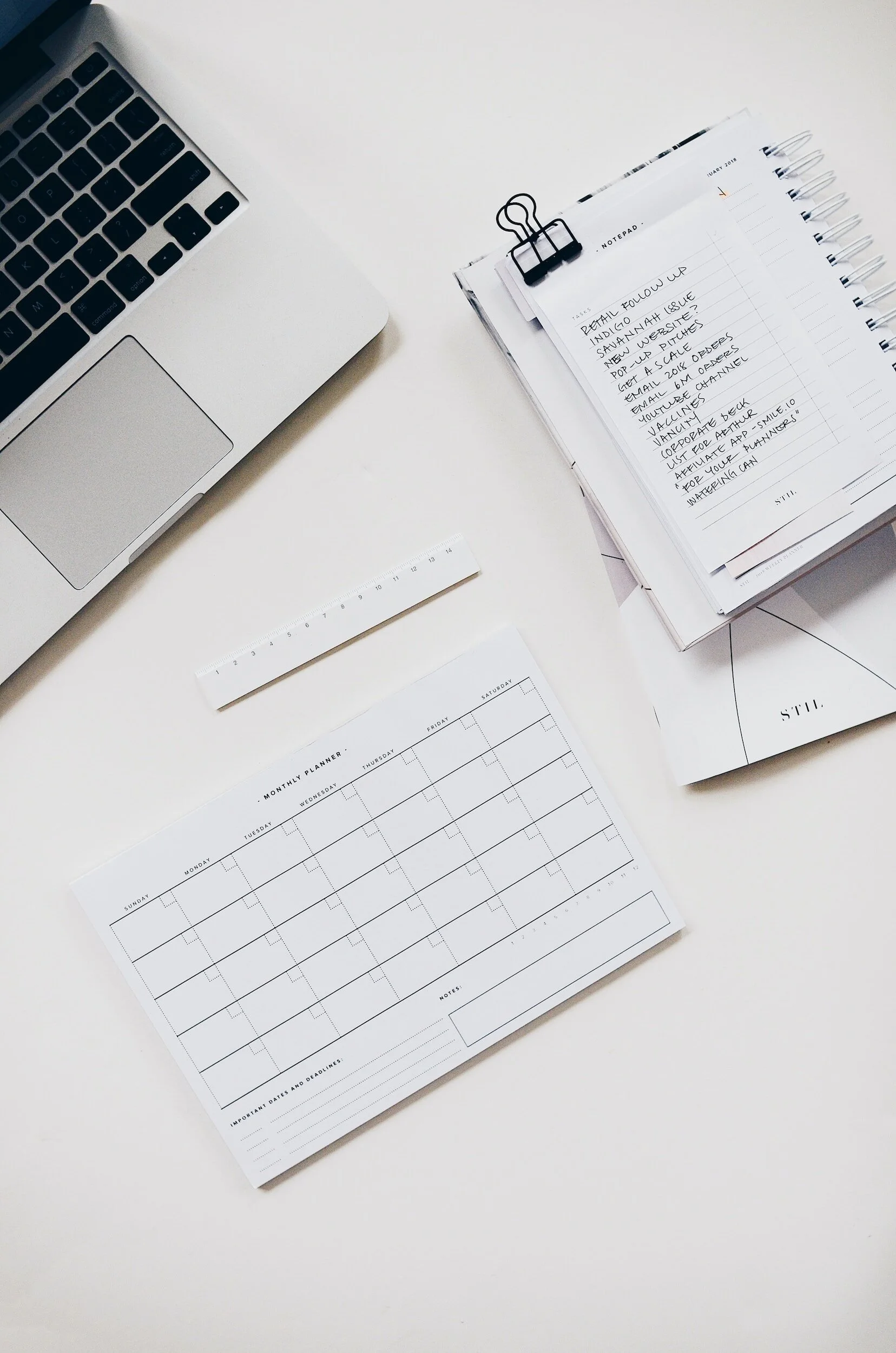 White desk with a silver laptop, a ruler, a monthly planner, a notepad with handwritten notes, and some papers.