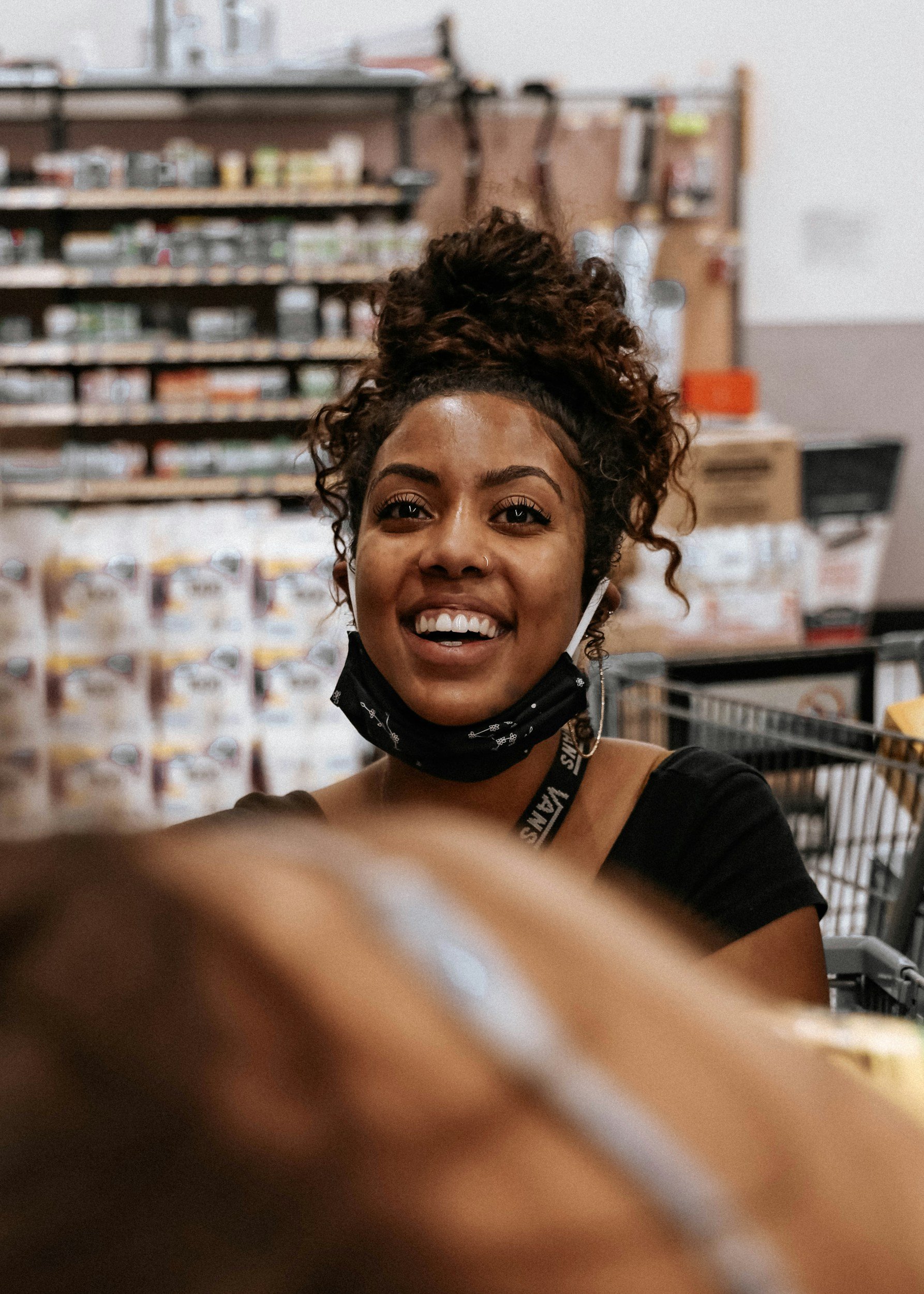 A young woman with curly hair in a bun, wearing a face mask around her neck, smiling in a store with shelves of products in the background.