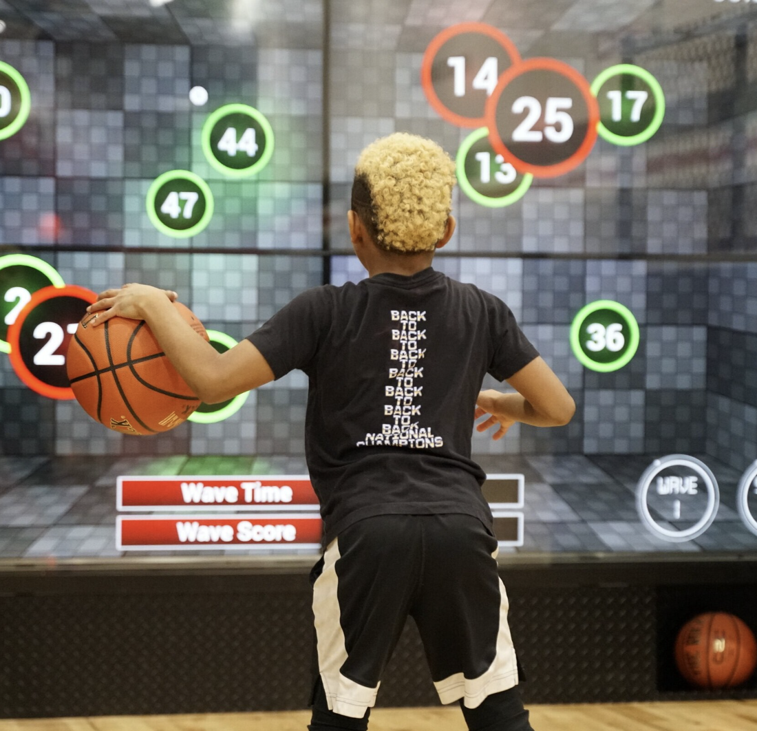 A young person with blonde curly hair holding a basketball, standing in front of a digital basketball arcade game screen with numbered circles and scoring prompts.