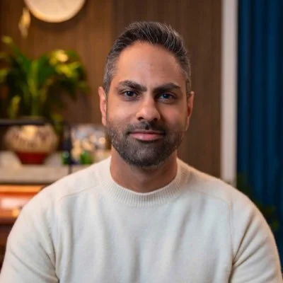 A man with short dark hair, beard, and mustache wearing a white sweater in a room with wooden walls, green plants, and a blue curtain.