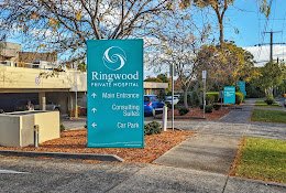 Sign for Ringwood Private Hospital indicating main entrance, consulting rooms, and car park, with trees and parked cars in the background.