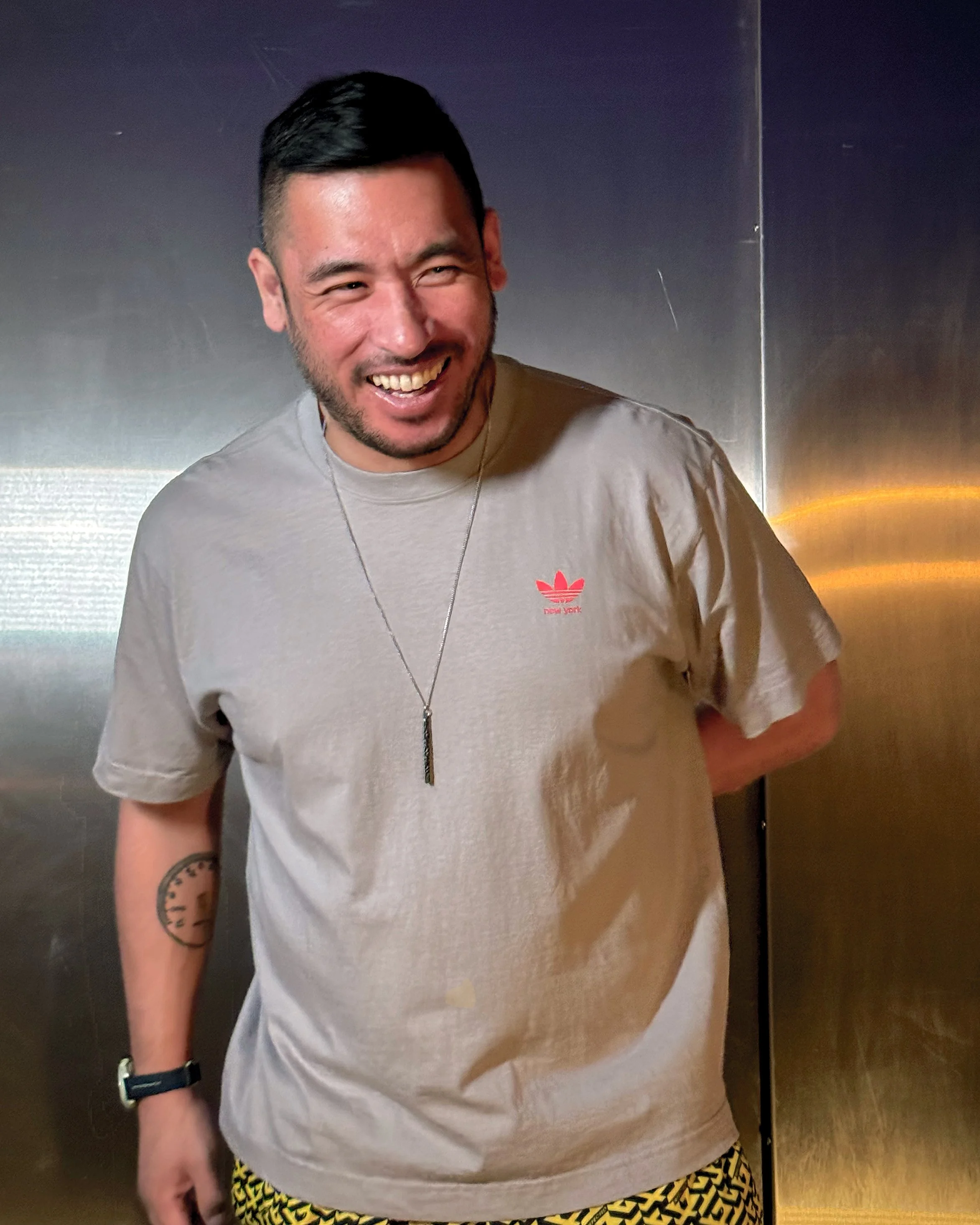 A smiling man with dark hair, wearing a gray T-shirt with a small red logo on the chest, and patterned yellow and black shorts. He has a tattoo on his left forearm, a necklace, a wristwatch, and an earpiece.