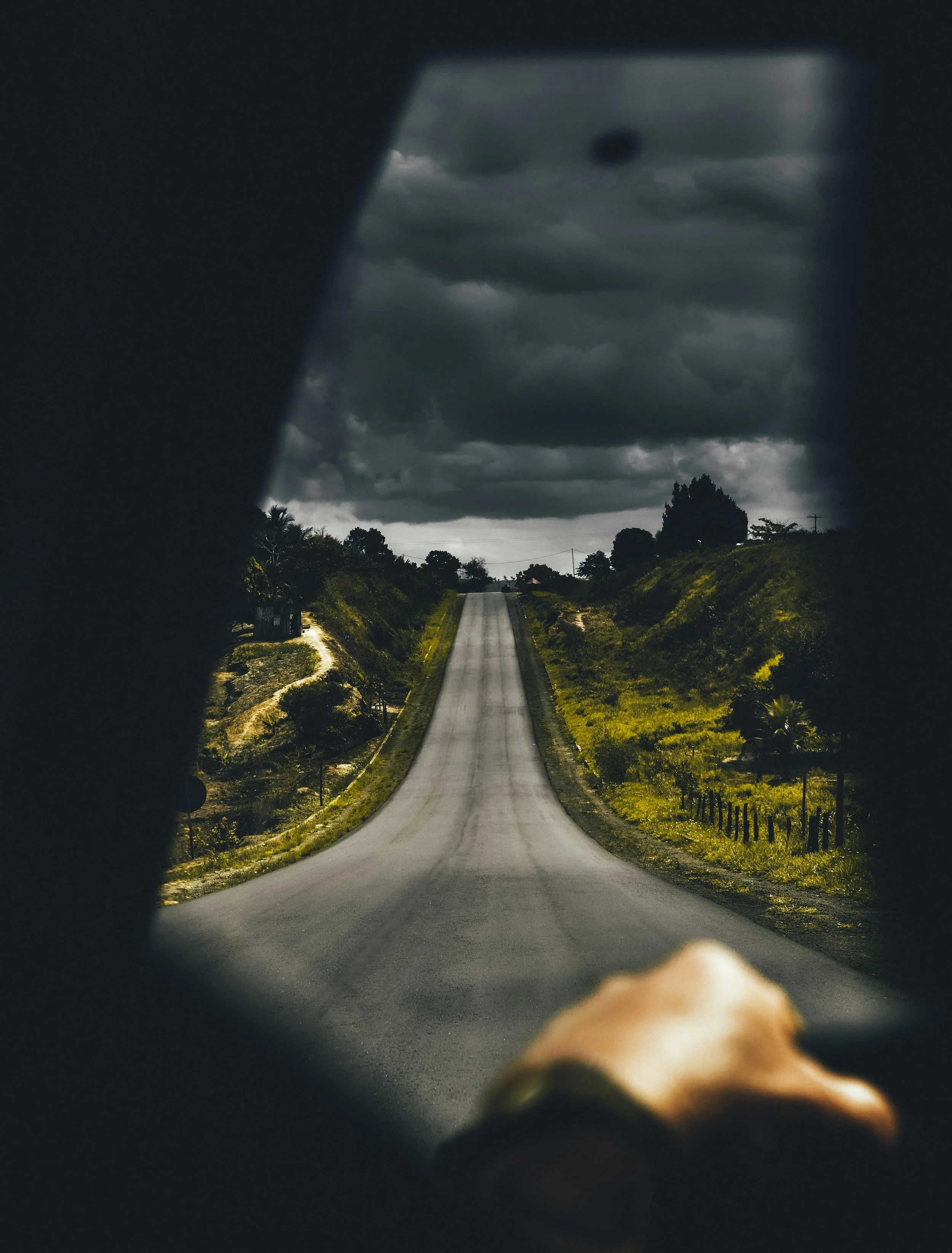 A long, empty road stretching into the distance under a dark, cloudy sky, viewed through a car's side mirror with part of the person's hand visible at the bottom.
