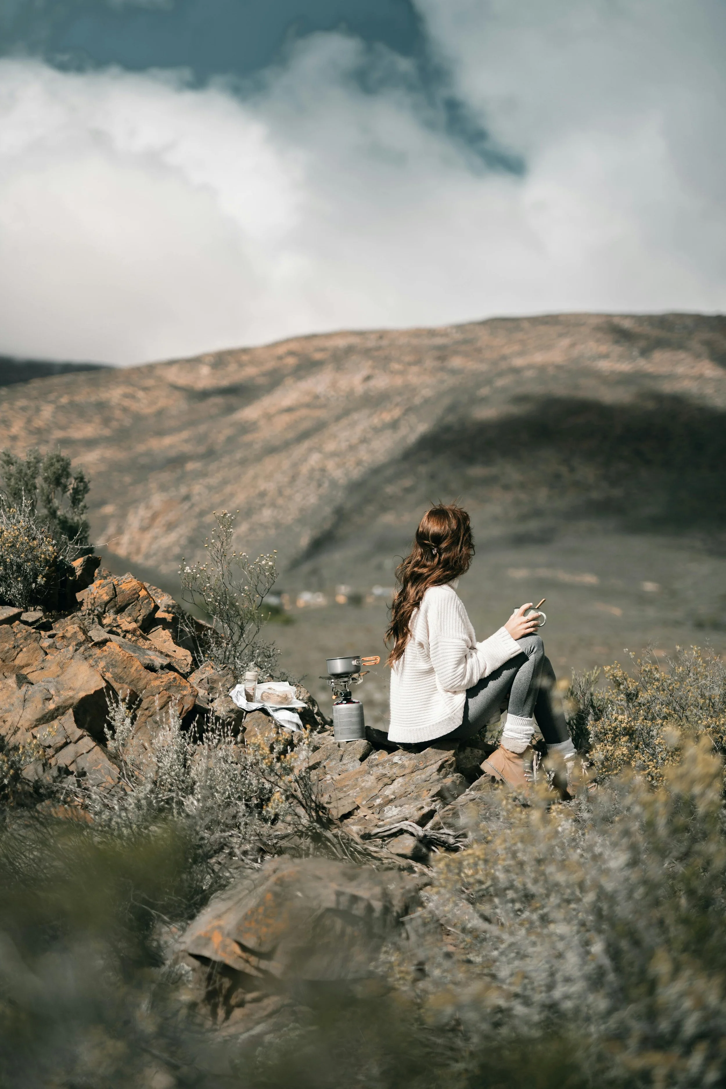 A woman with long brown hair sitting on rocks in a mountainous landscape, looking at her phone, with a camping stove and supplies nearby.