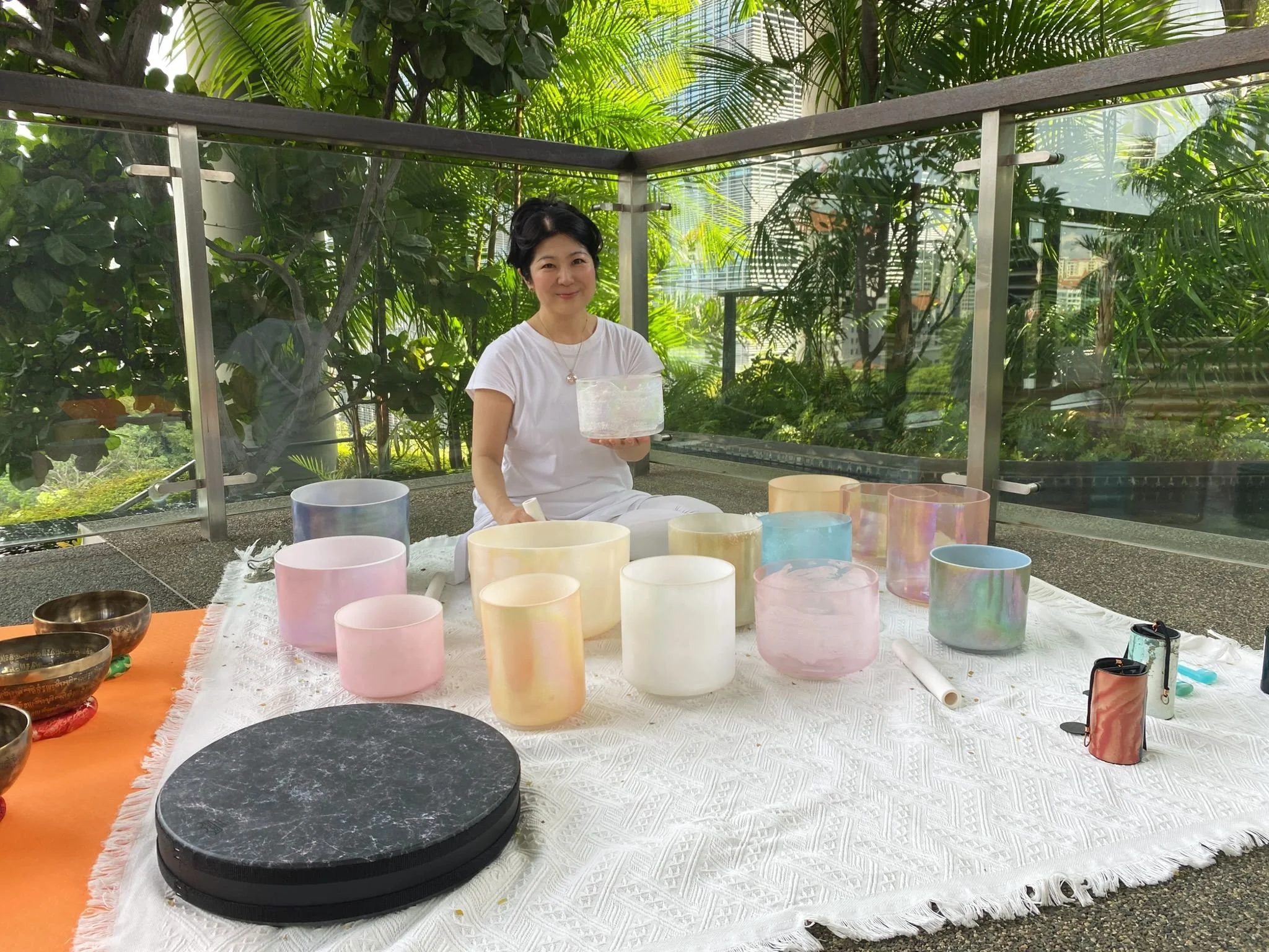 A woman in a white outfit sitting on a blanket surrounded by colorful singing bowls on a rooftop with greenery in the background, holding a clear bowl.