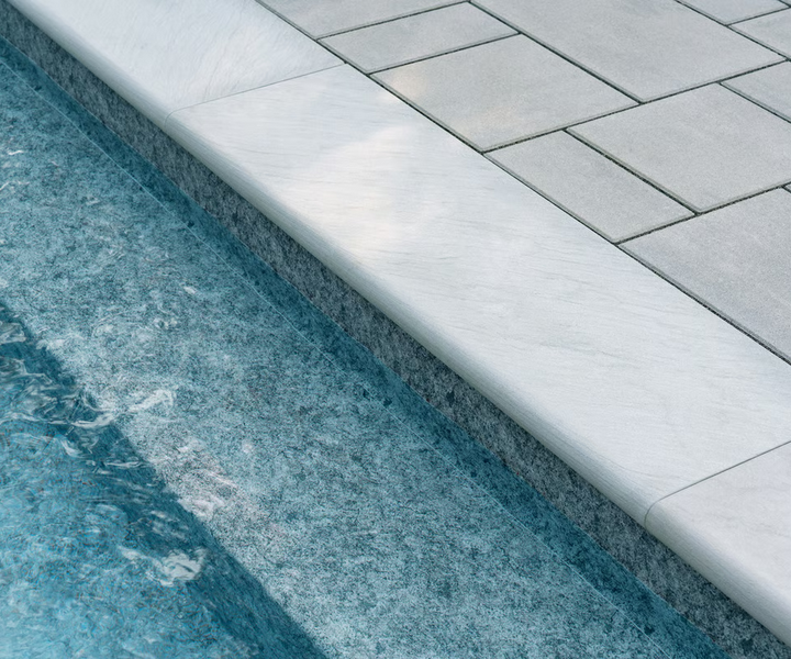 Close-up of a pool's edge with a white marble coping and gray paving stones, next to blue water.