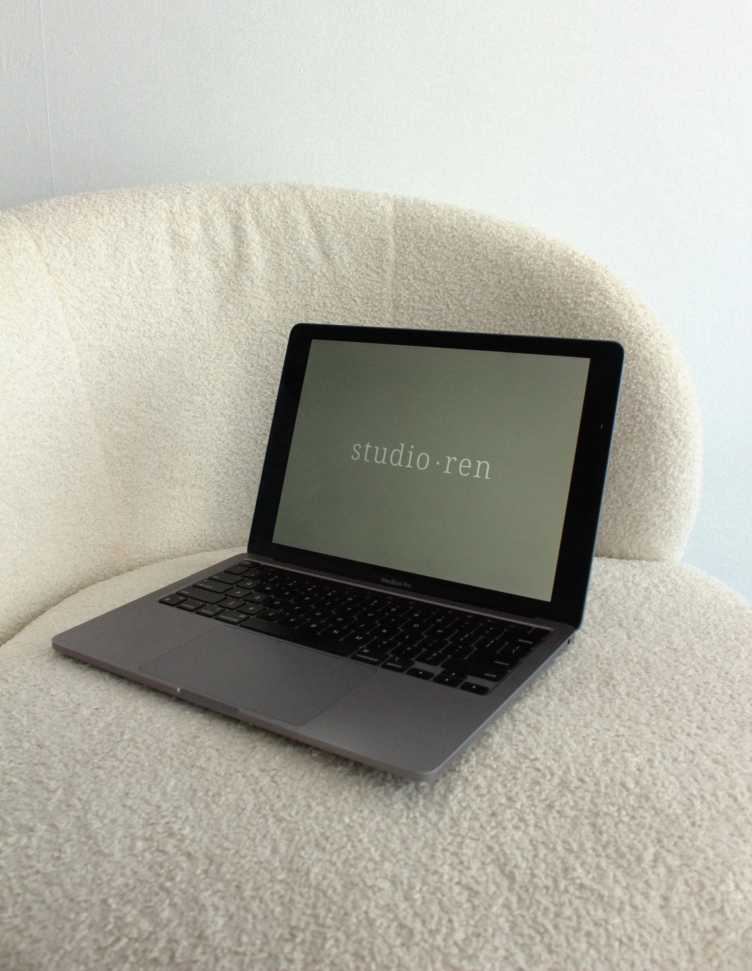 A silver MacBook Pro with a black keyboard on a cream-colored textured chair, displaying the text 'studio.ren' on its screen.