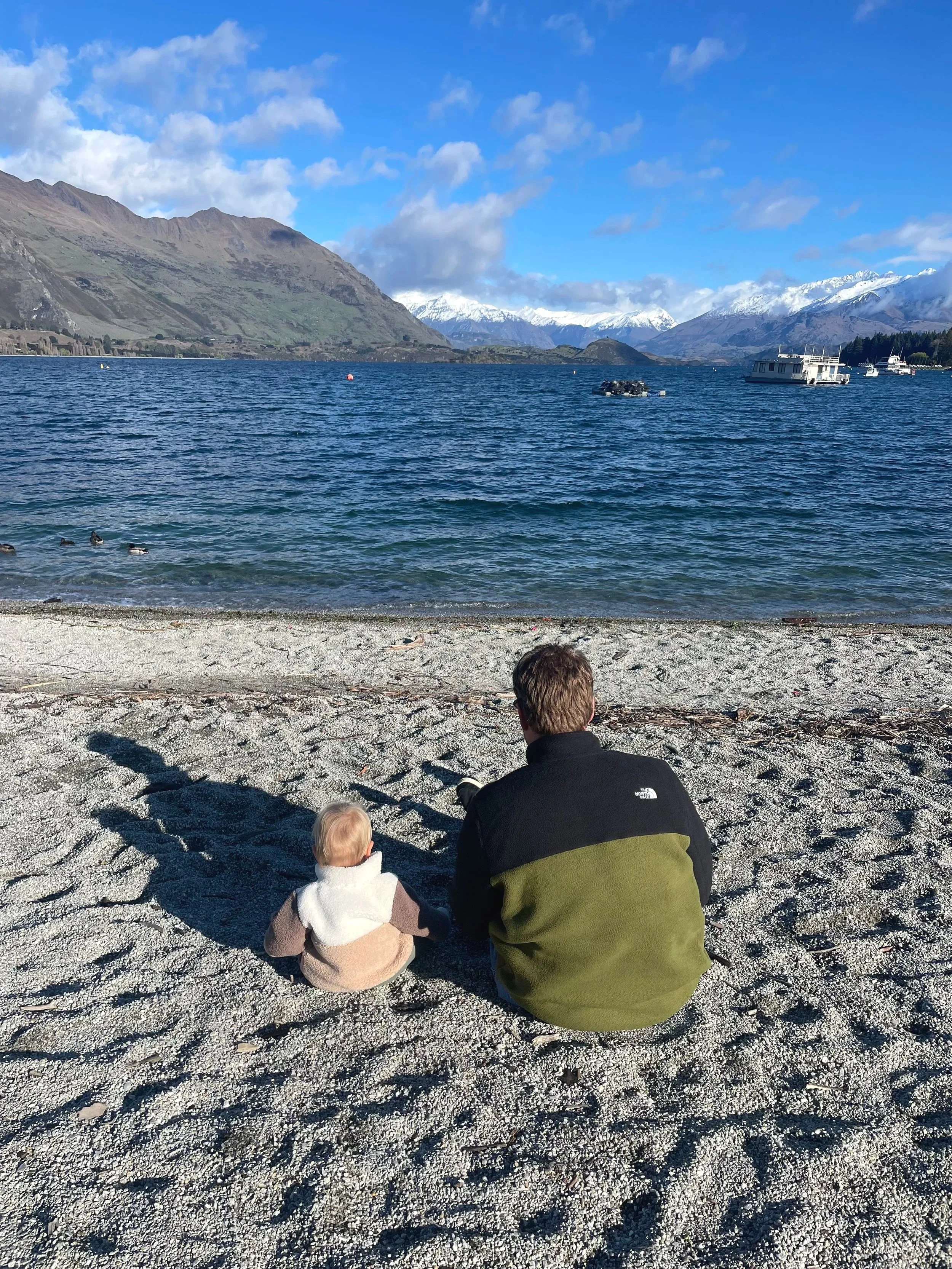 A man and a small child sitting on a rocky beach, facing a large lake with mountains and snowy peaks in the background, under a partly cloudy sky.