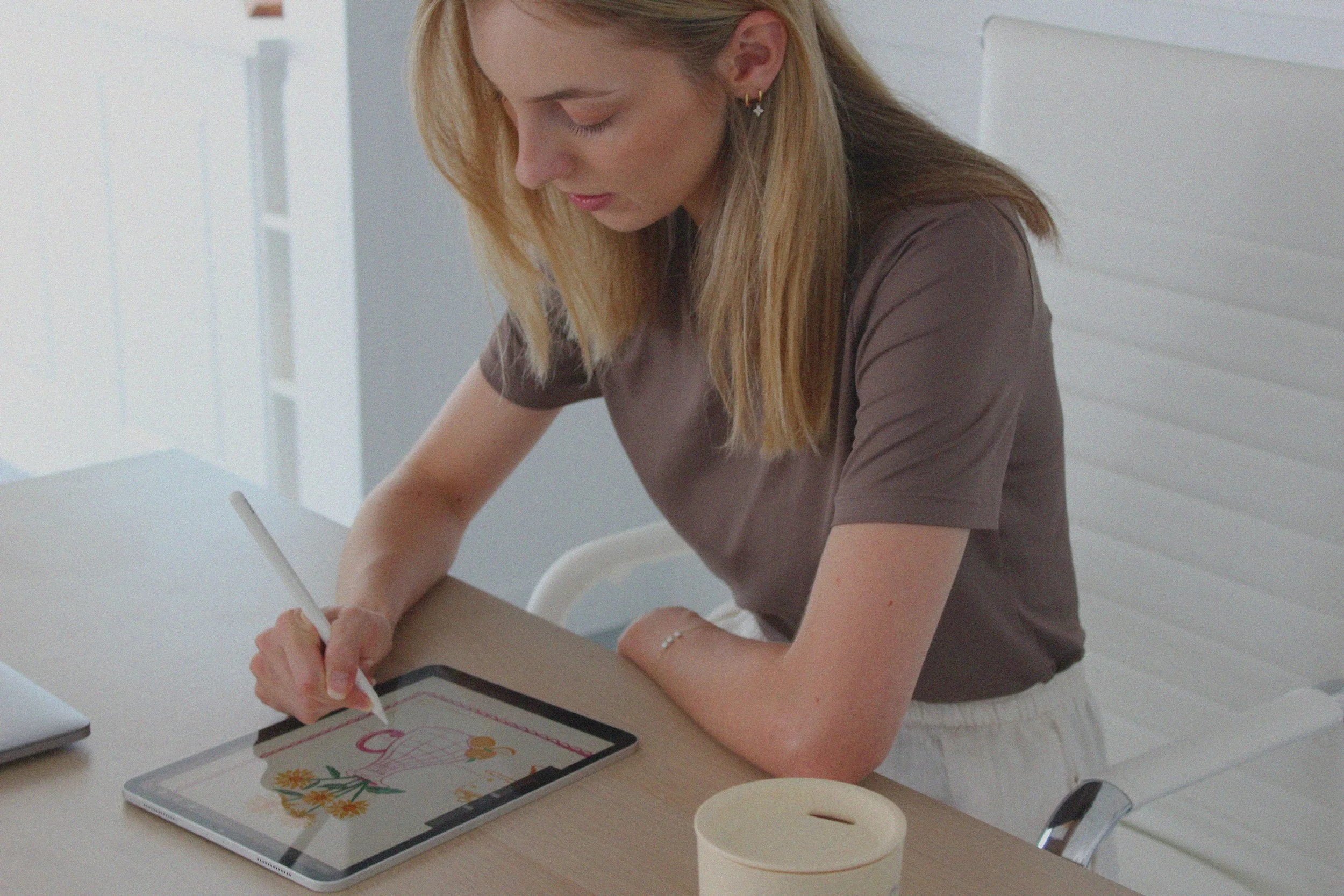 A young woman with blonde hair is drawing on an iPad. She is sitting at a light-coloured table in a bright room, with a notepad and a coffee cup on the table.