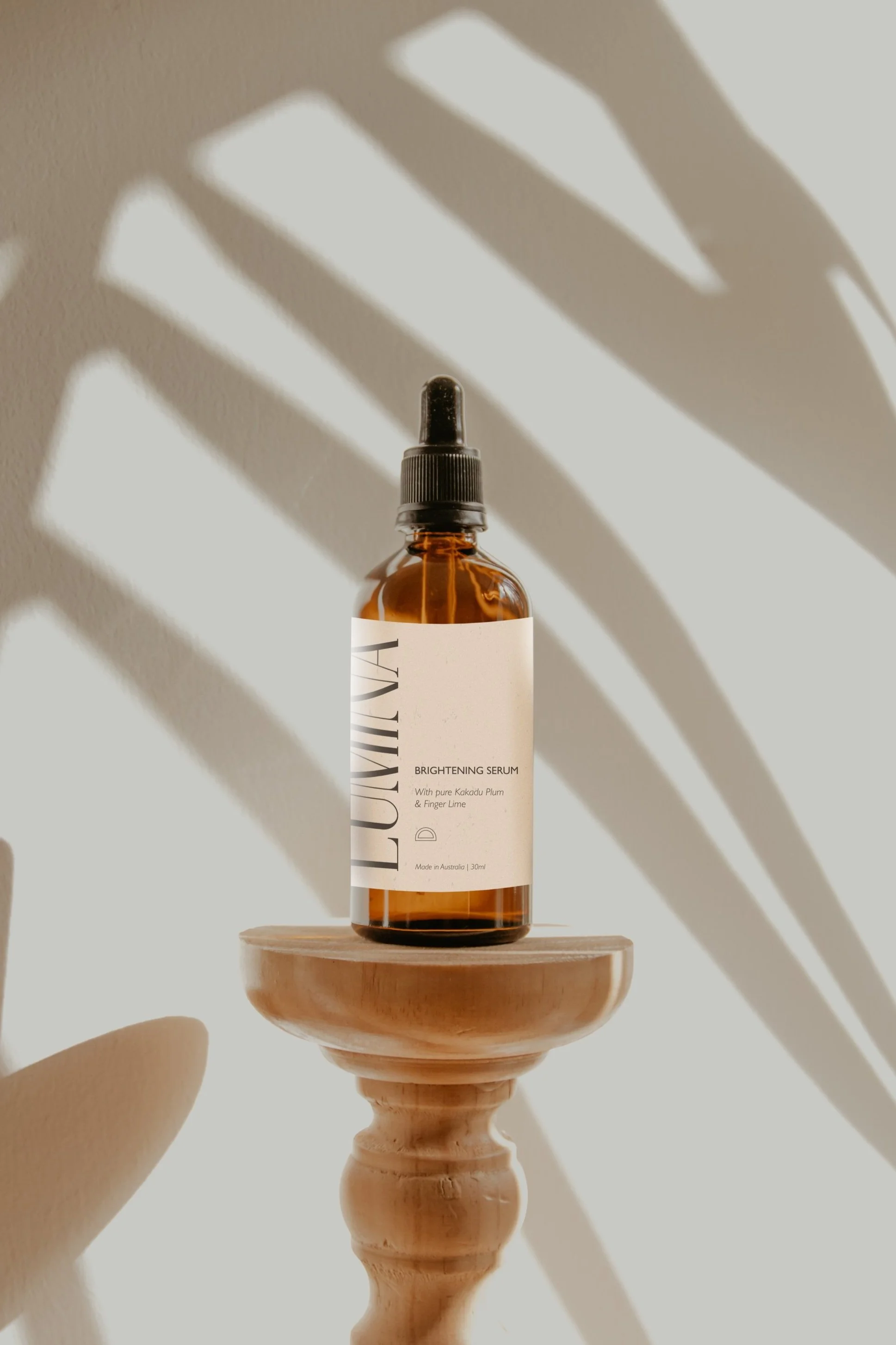 A brown glass dropper bottle labeled "LUMIN" with a white label that reads "Brightening Serum" and mentions ingredients like pure Kakadu plum and finger lime. The bottle is placed on a wooden pedestal with shadows of leaves cast on the wall behind it.