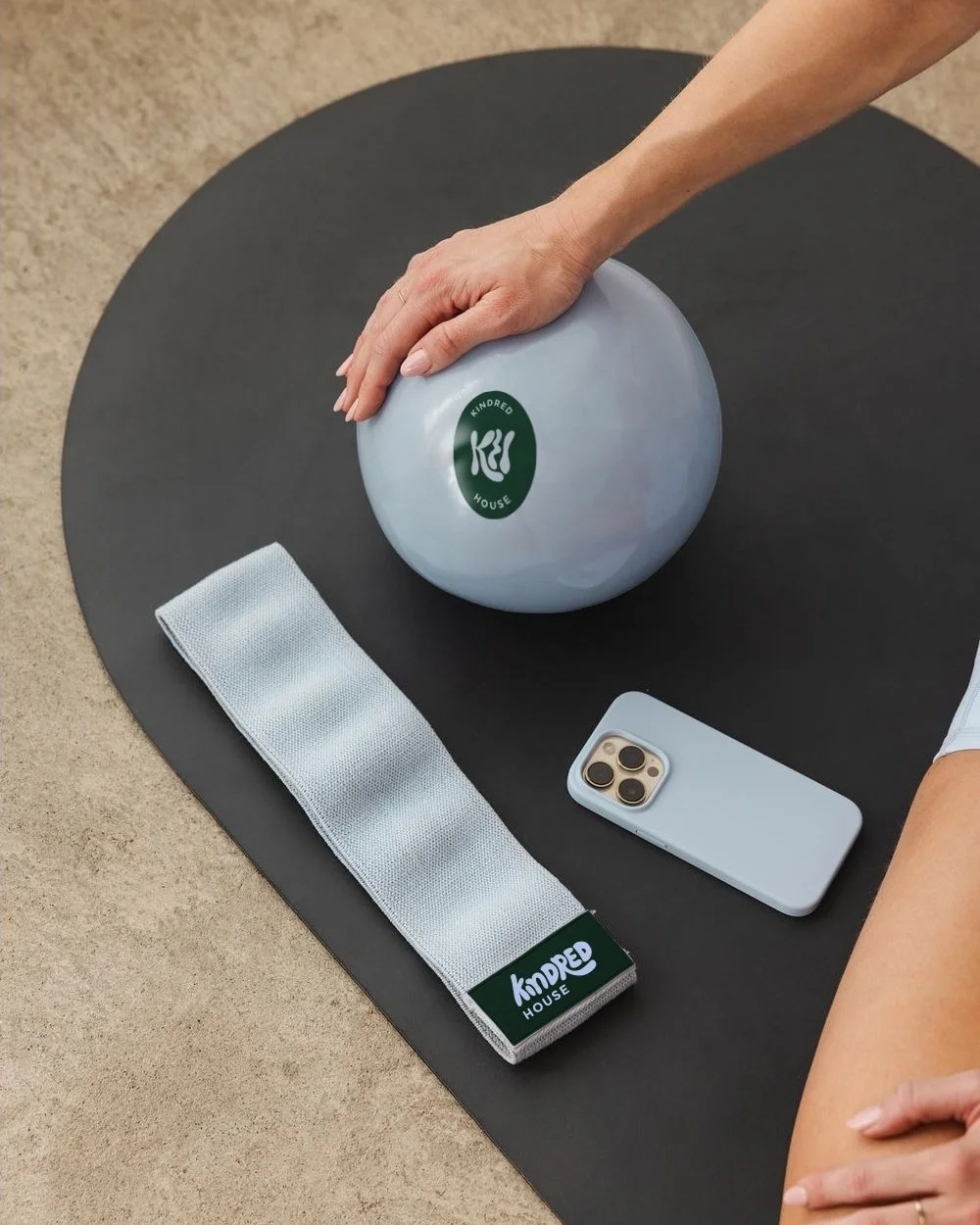 A hand resting on a gray exercise ball with a green logo that says 'Kindred House,' next to a white fabric belt with a similar logo, a smartphone with a light gray case, and a black yoga mat, all on a light-colored floor.