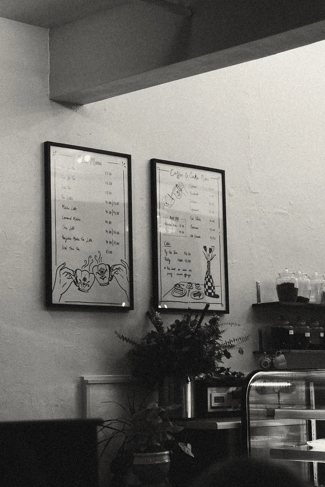 Black and white photo of a cafe interior with two framed menu boards on the wall, a potted plant below the boards, and a display case on the right. The menus feature illustrations of hands holding cups and a vase with flowers.