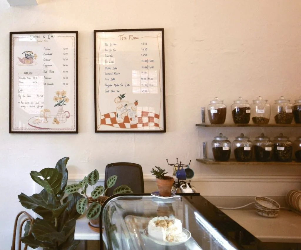 Cafe interior with coffee and tea menu boards on the wall, jars of coffee beans on shelves, plants, and a display case with baked goods.