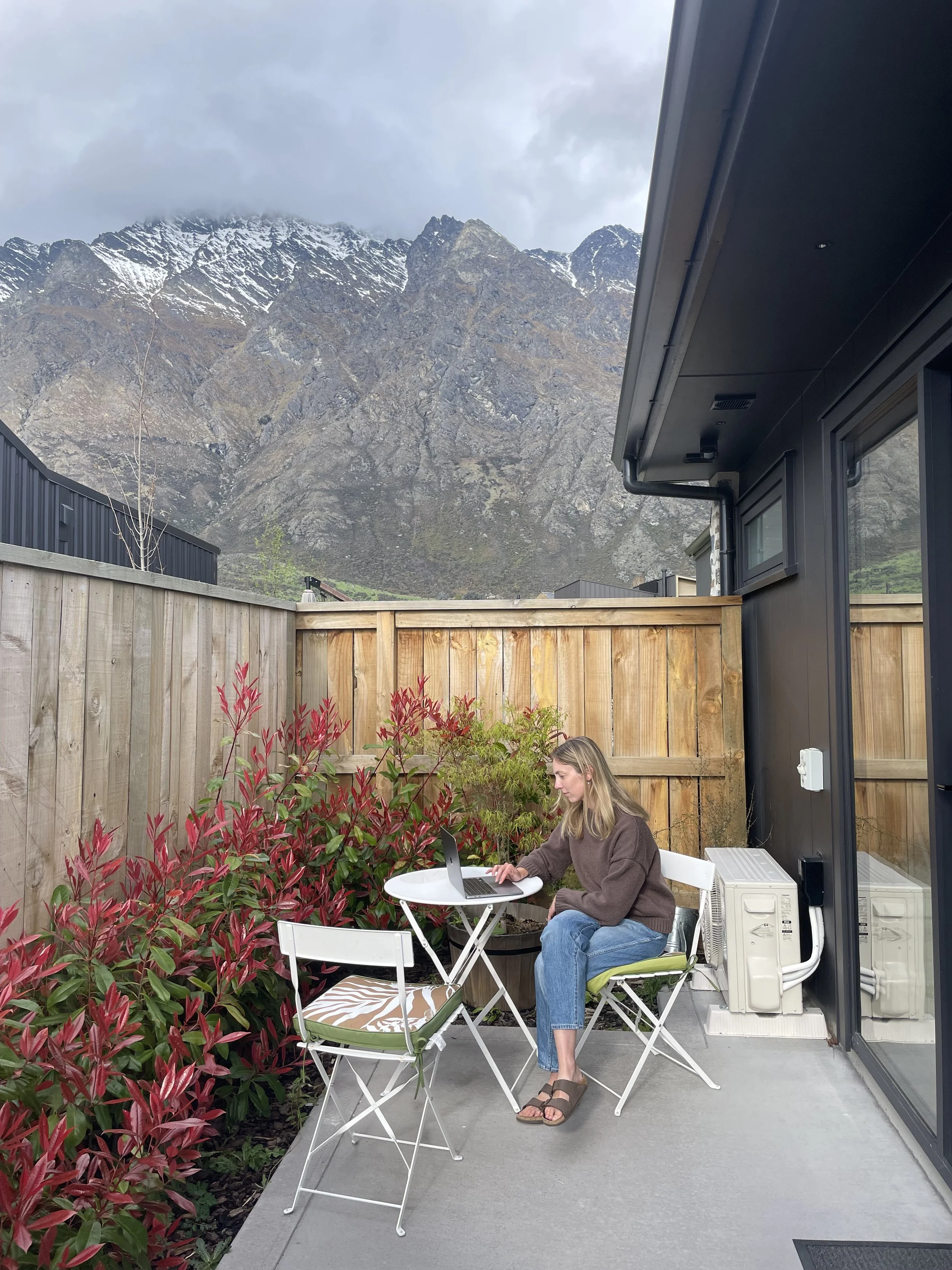 A woman sitting at a small white table on a balcony with a laptop, surrounded by red and green plants, with a mountain range in the background.