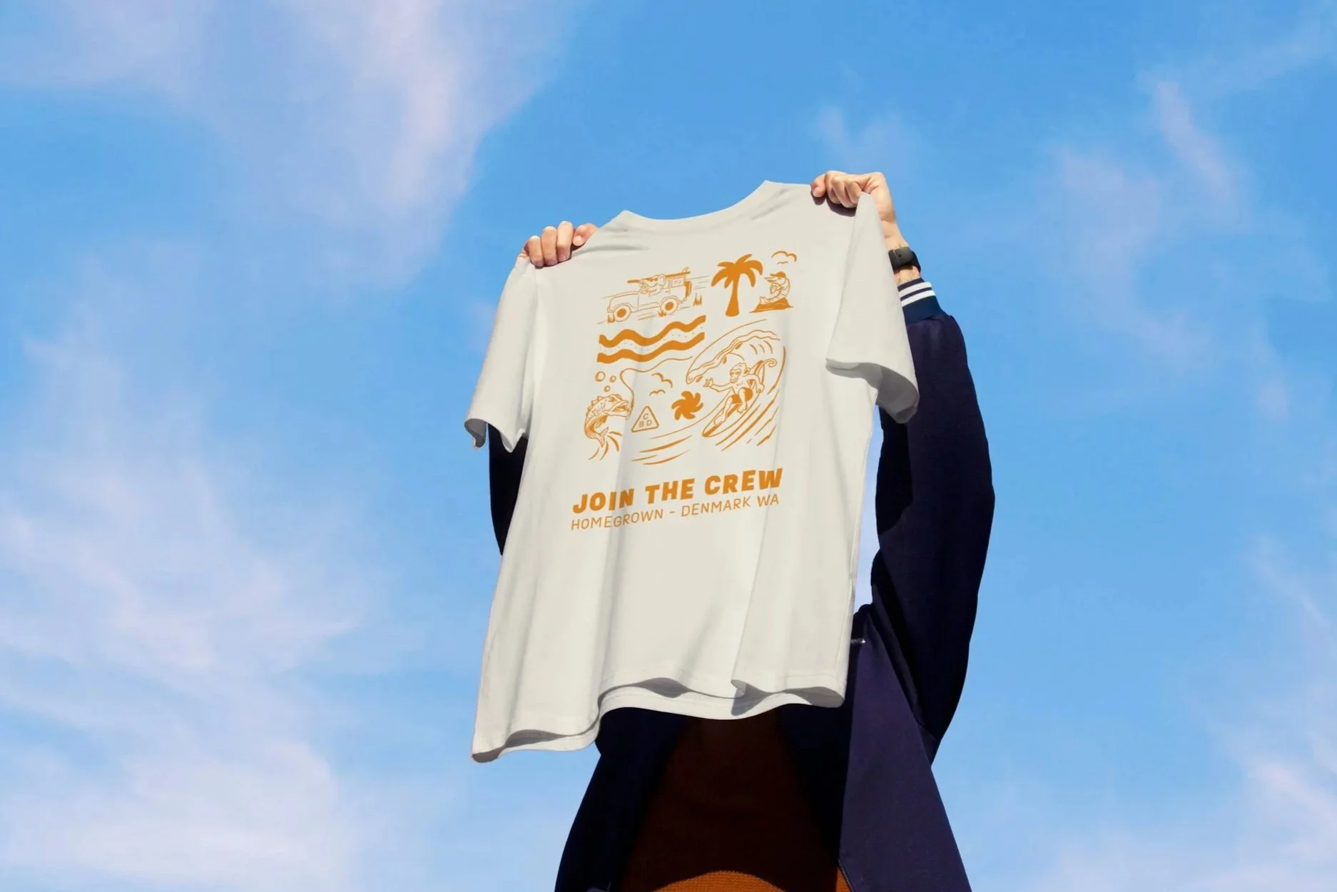 Person holding a white T-shirt with orange graphic and text against a blue sky. The graphic features a boat, palm trees, waves, a surfer, and the text "JOIN THE CREW HOMETOWN - DENMARK WA."