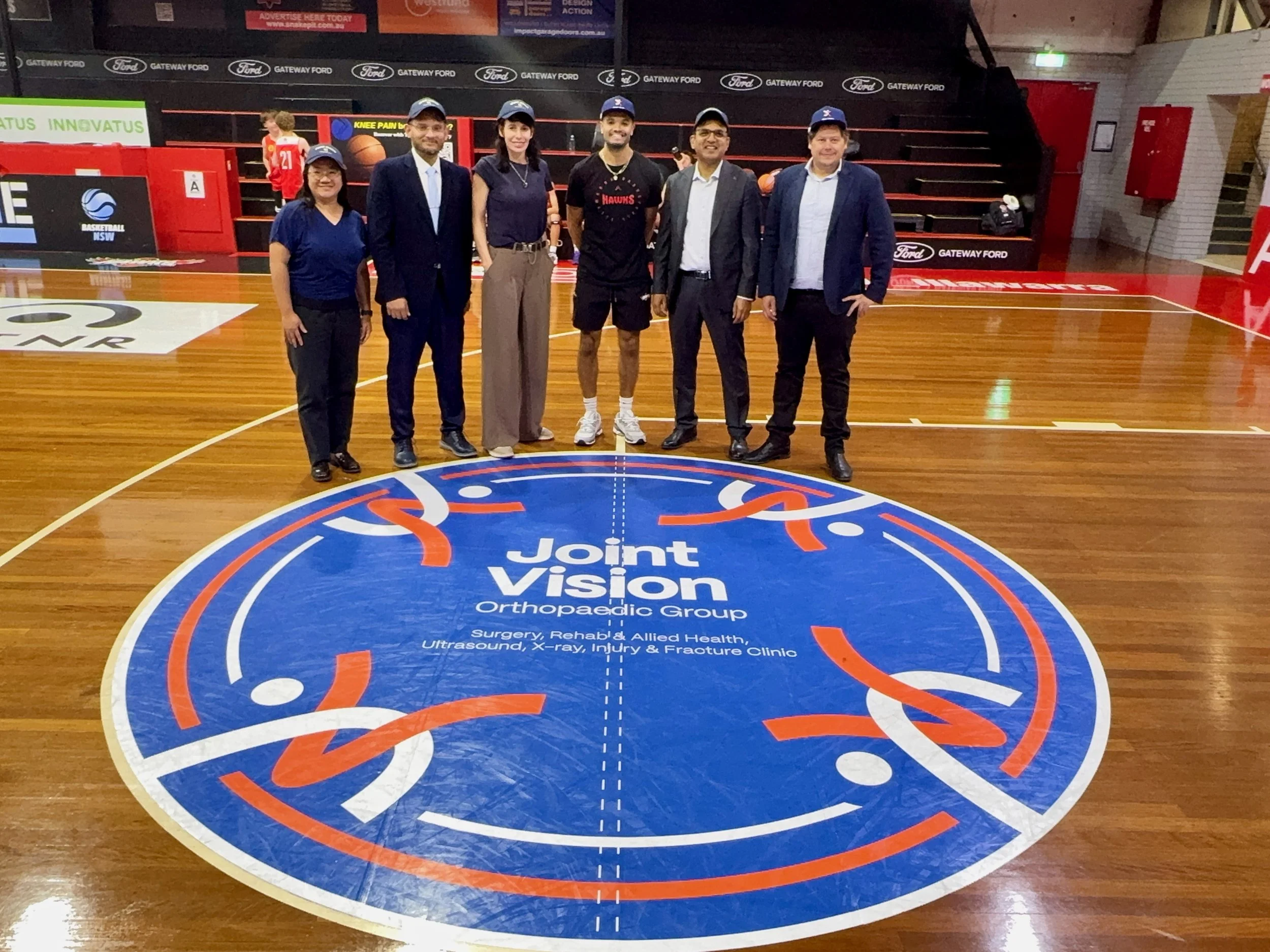 Joint Vision Orthopaedic Group team with Illawarra Basketball Association representatives standing on centre court logo at Snakepit during partnership launch