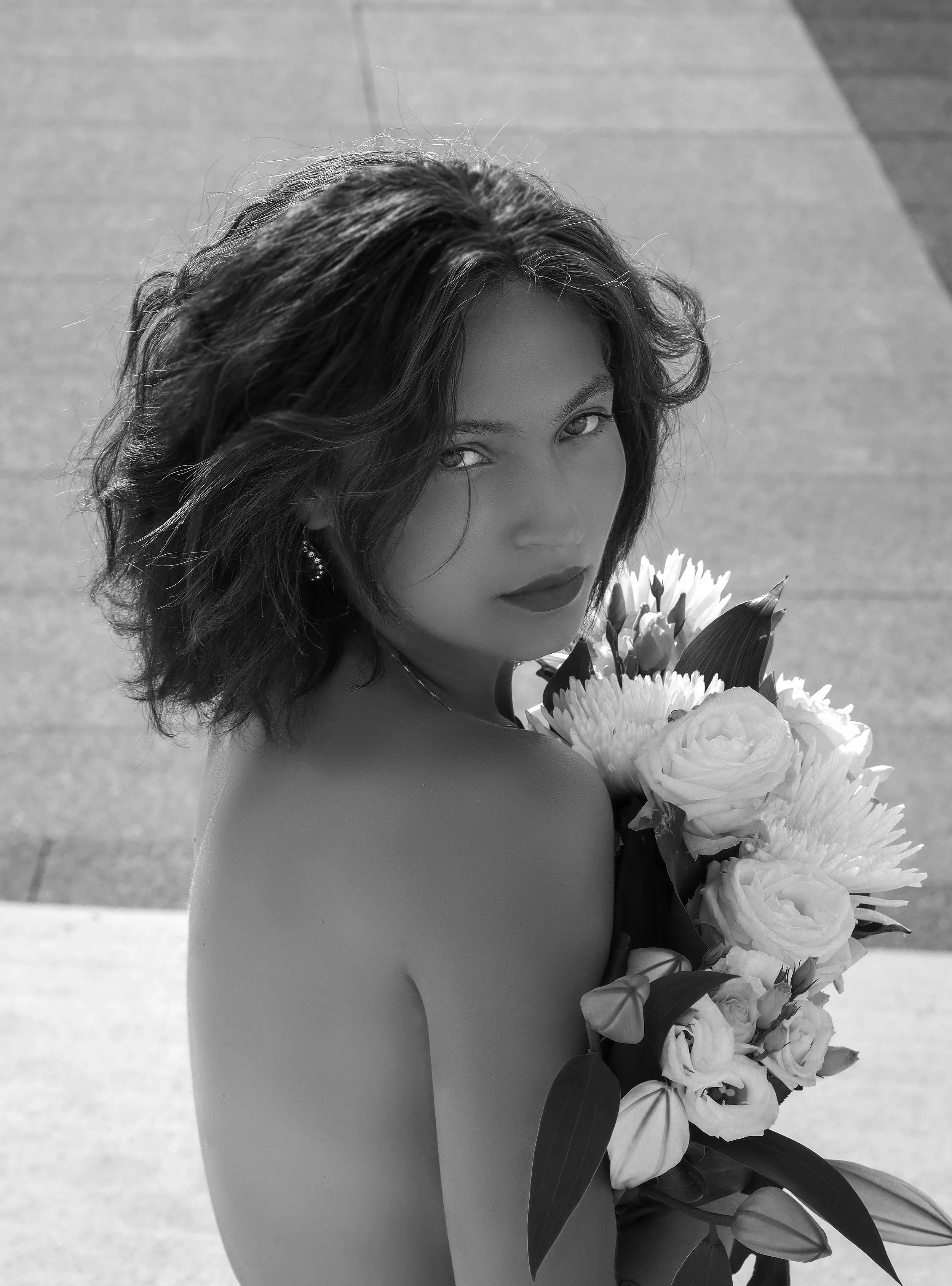 Black and white photo of a woman with wavy hair holding a large bouquet of flowers, looking directly at the camera.
