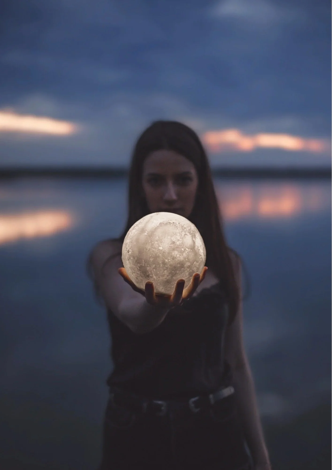 A woman holding a glowing moon in her outstretched hand during dusk or evening.