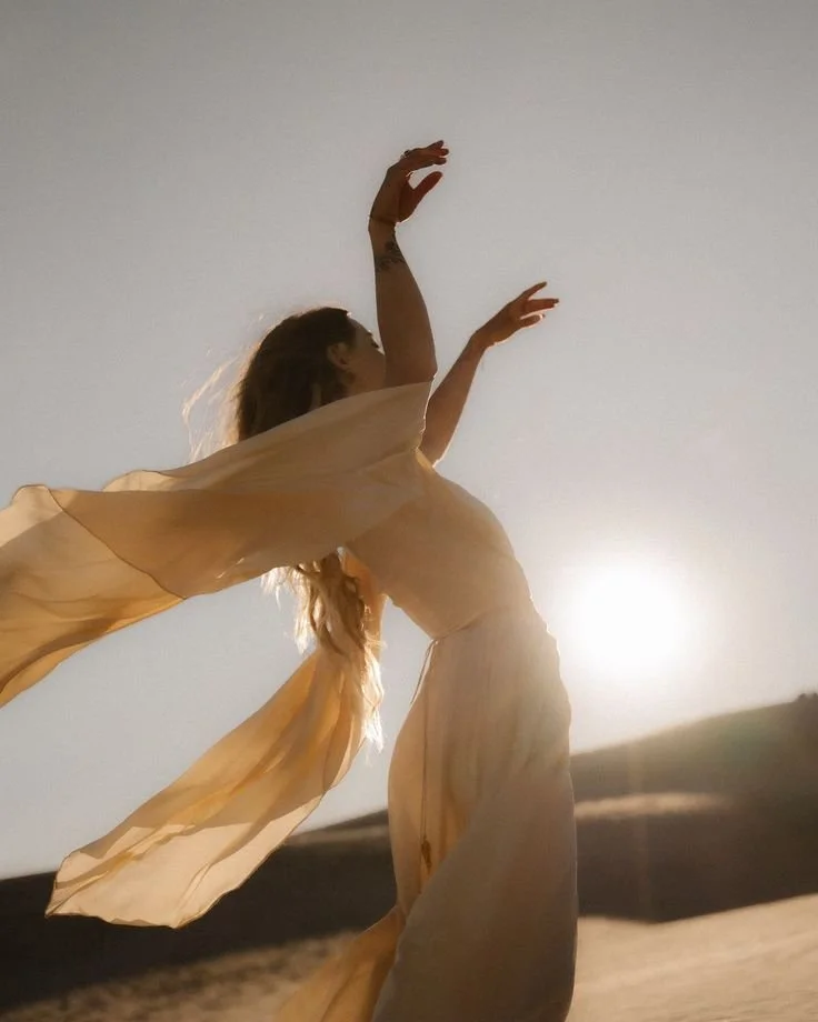 A woman dancing or posing outdoors at sunset, wearing a flowing, light-colored dress with her arms raised upward.