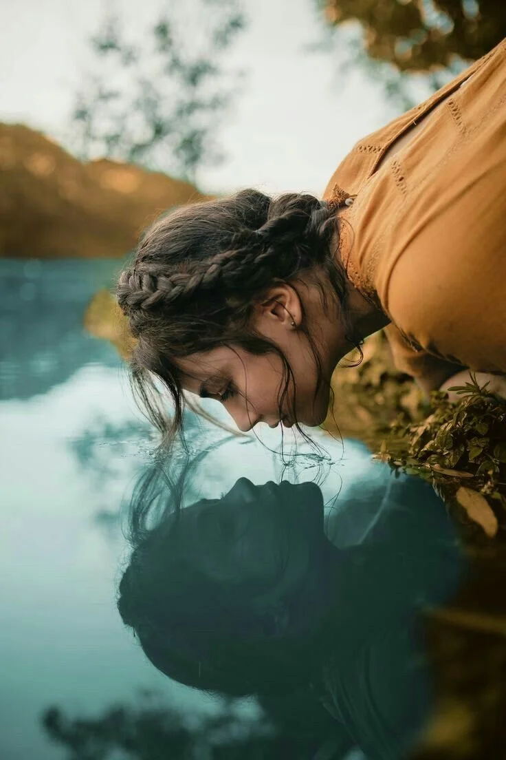 A woman with braided hair and earrings leaning over and touching her face to the water of a lake, creating a reflection of her face in the water.