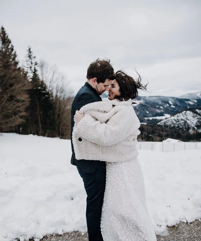 Winterhochzeit in den Alpen beim Ansitz Wartenfels fotografiert. Die frisch vermählten stehen vom Schnee umgeben vor der Kamere´a der Fotografin Claudia Weaver, Sie lächeln und stehen sich gegenüber.