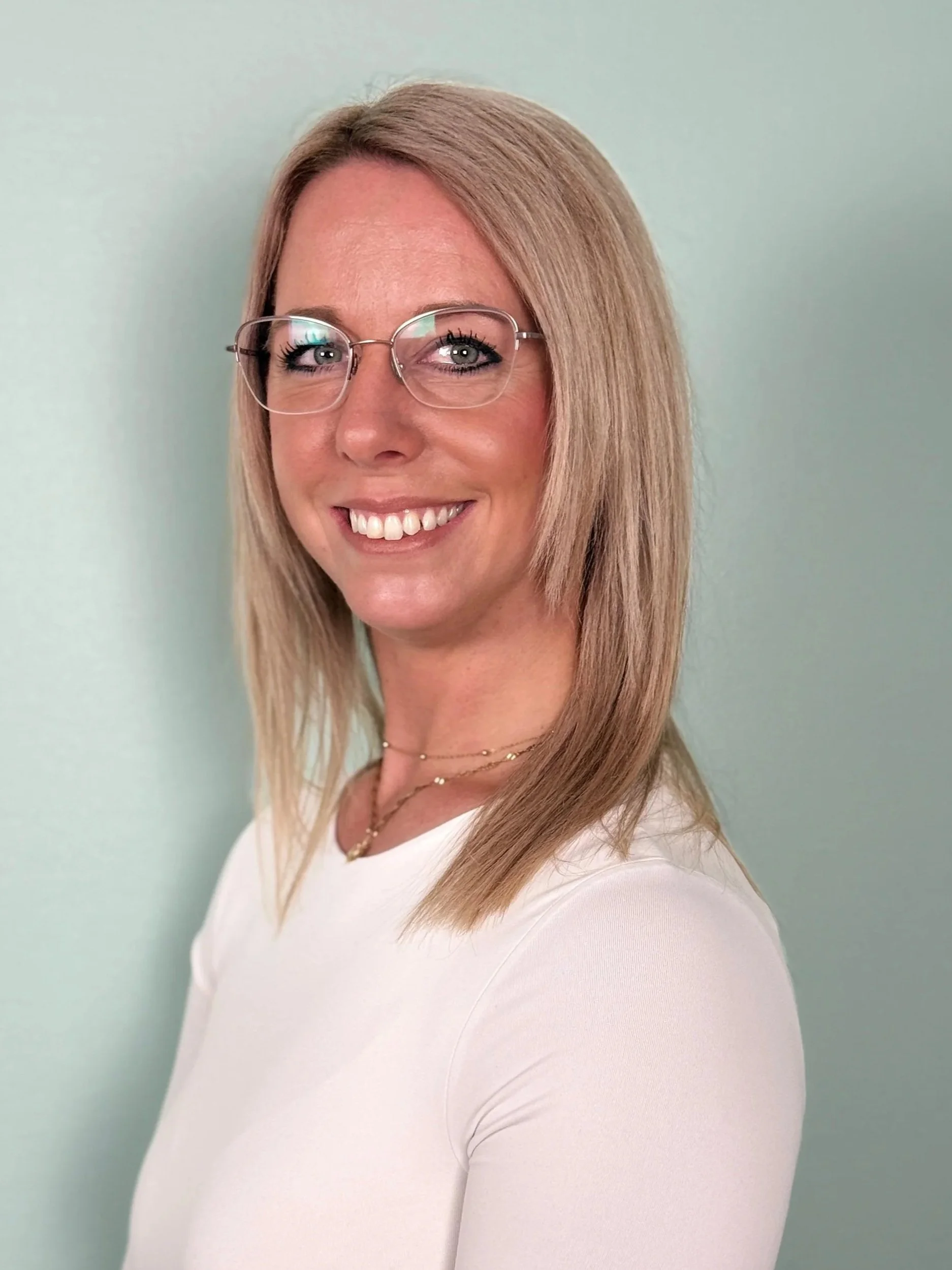 Eine lachende Frau mit brünettem, geraden Haar, trägt Brillen mit dünnem Rahmen und eine weiße Bluse, vor einem hellen Hintergrund.