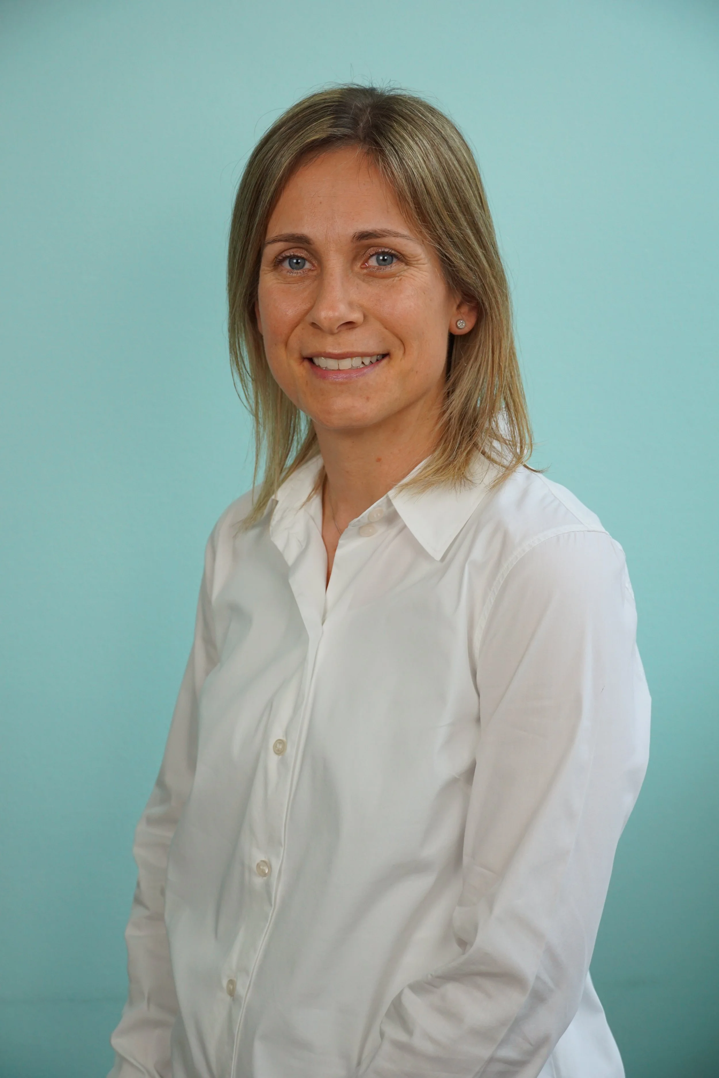 Porträt einer lachenden Frau mit blonden Haaren, weißem Hemd vor einem hellblauen Hintergrund.