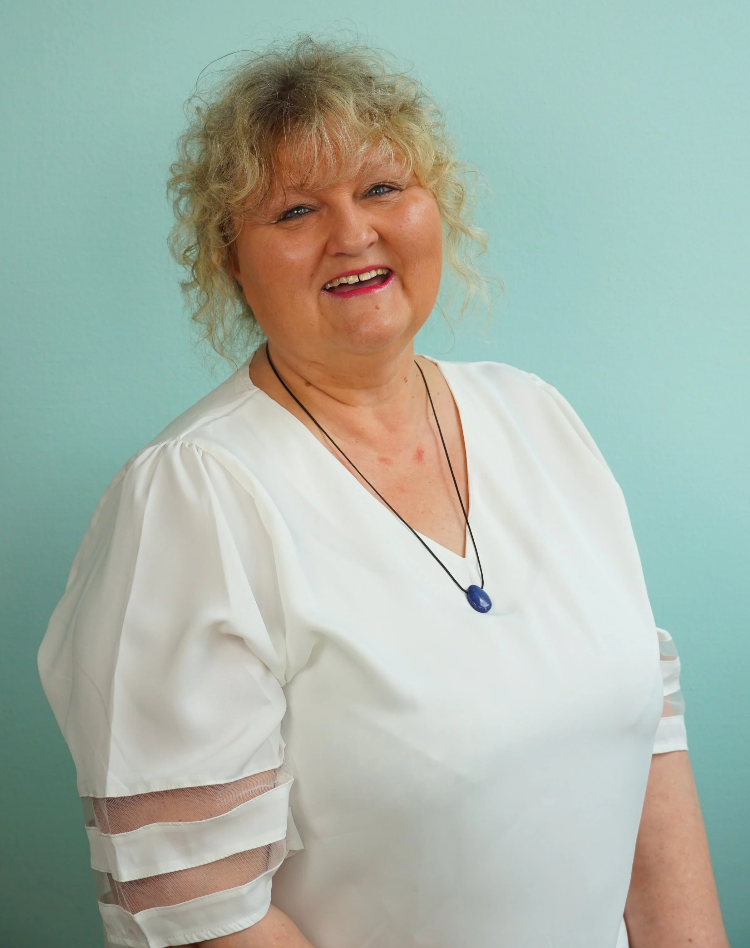 Eine lachende ältere Frau mit kurzen, lockigen, blonden Haaren, trägt eine weiße Bluse mit durchsichtigen Streifen an den Ärmeln, und ein schwarzes Halsband mit einem blauen Anhänger, vor einer mintgrünen Wand.