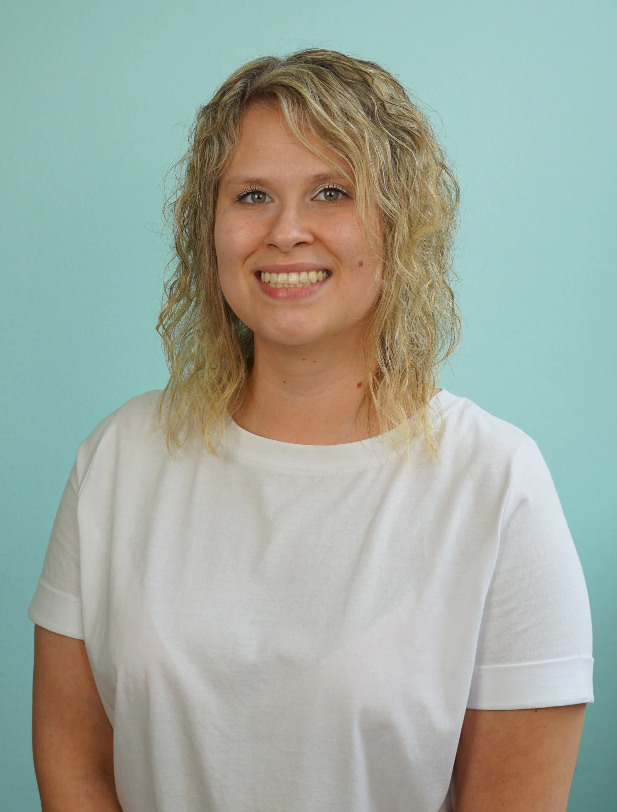 Ein junges Mädchen mit lockigem, blondem Haar vor einem hellblauen Hintergrund, lächelnd in die Kamera schauend.