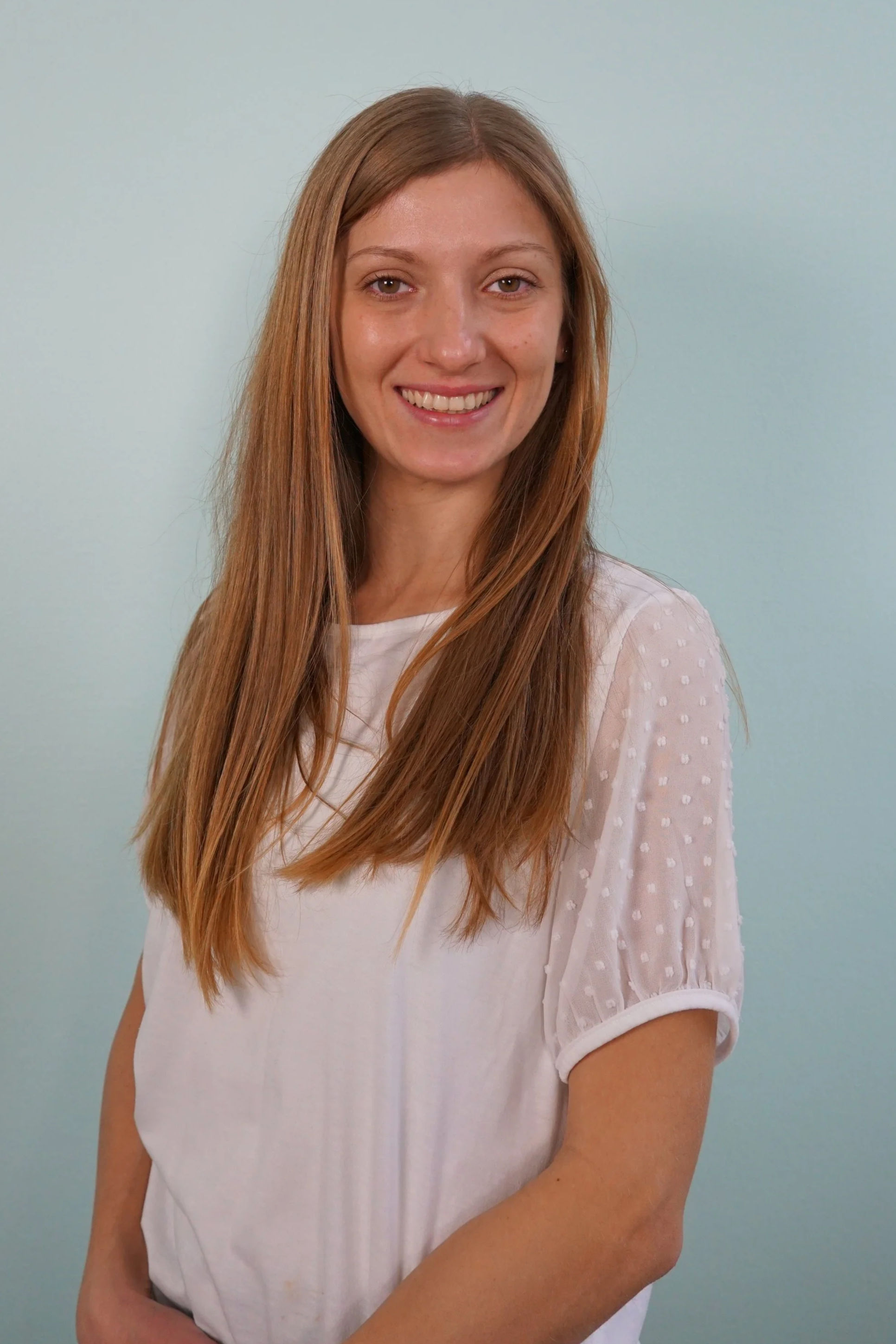 Ein lächelndes junges Mädchen mit langen, glatten, braunen Haaren, trägt ein weißes T-Shirt mit puffigen, transparenten Ärmeln, vor einem hellblauen Hintergrund.