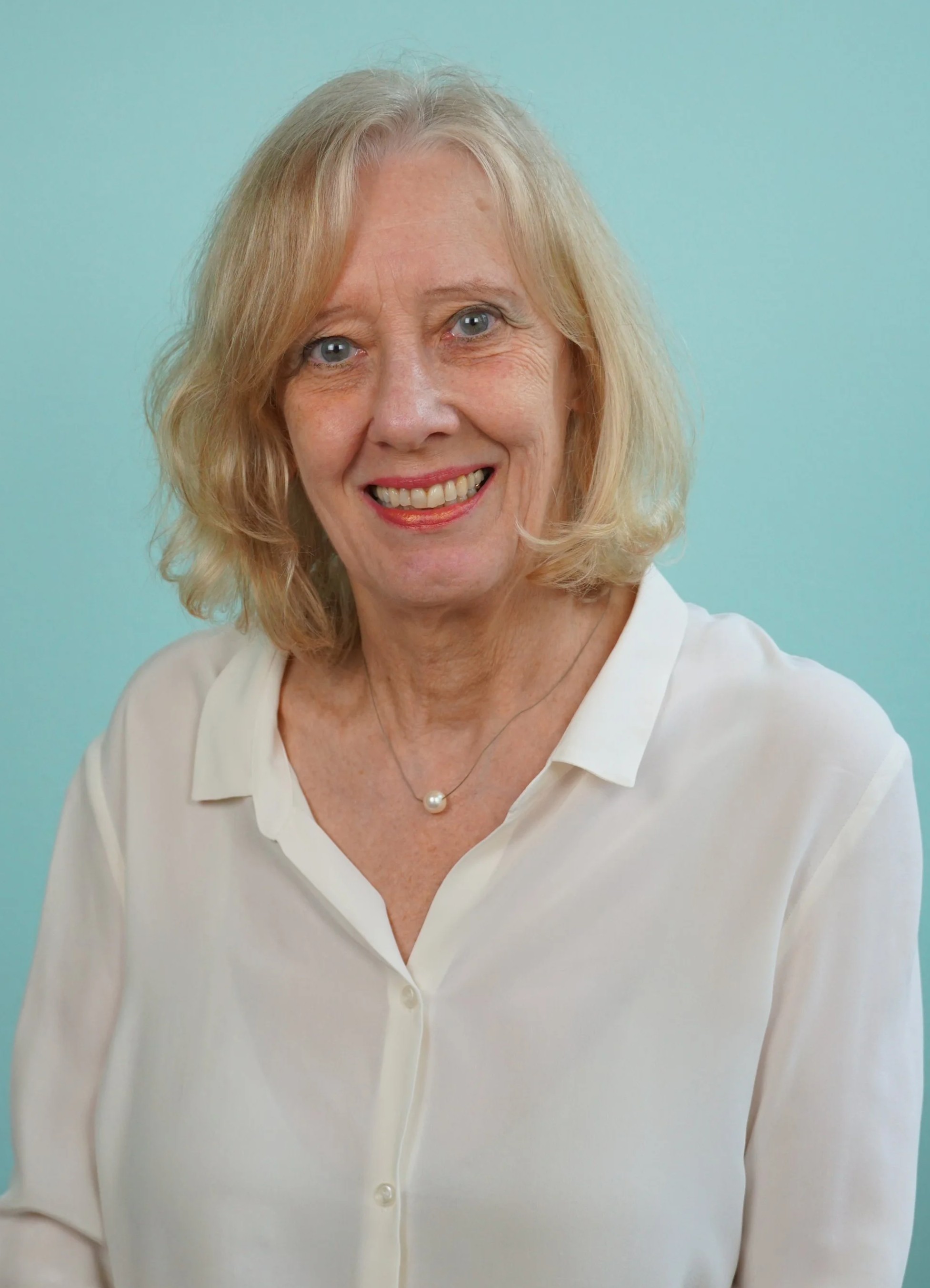 Porträt einer älteren Frau mit blonden, lockigen Haaren, die weiße Bluse trägt, vor einem hellblauen Hintergrund.