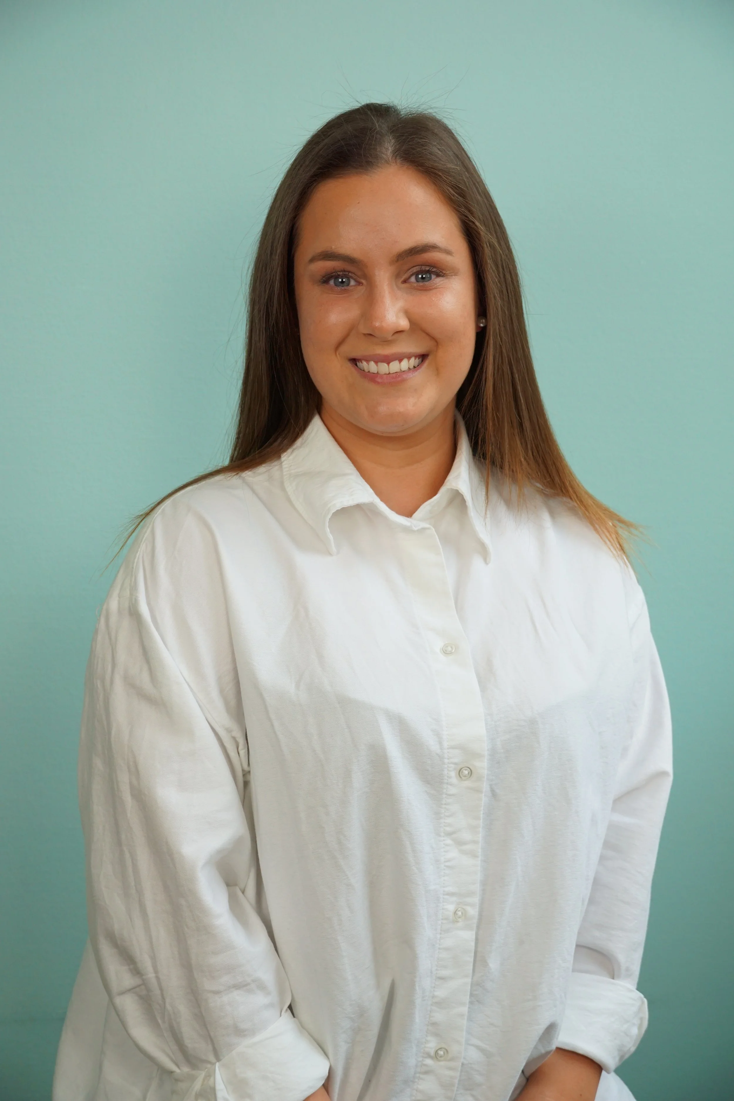 Junge Frau mit braunen Haaren, weißem Hemd, lächelnd, vor hellem Hintergrund.