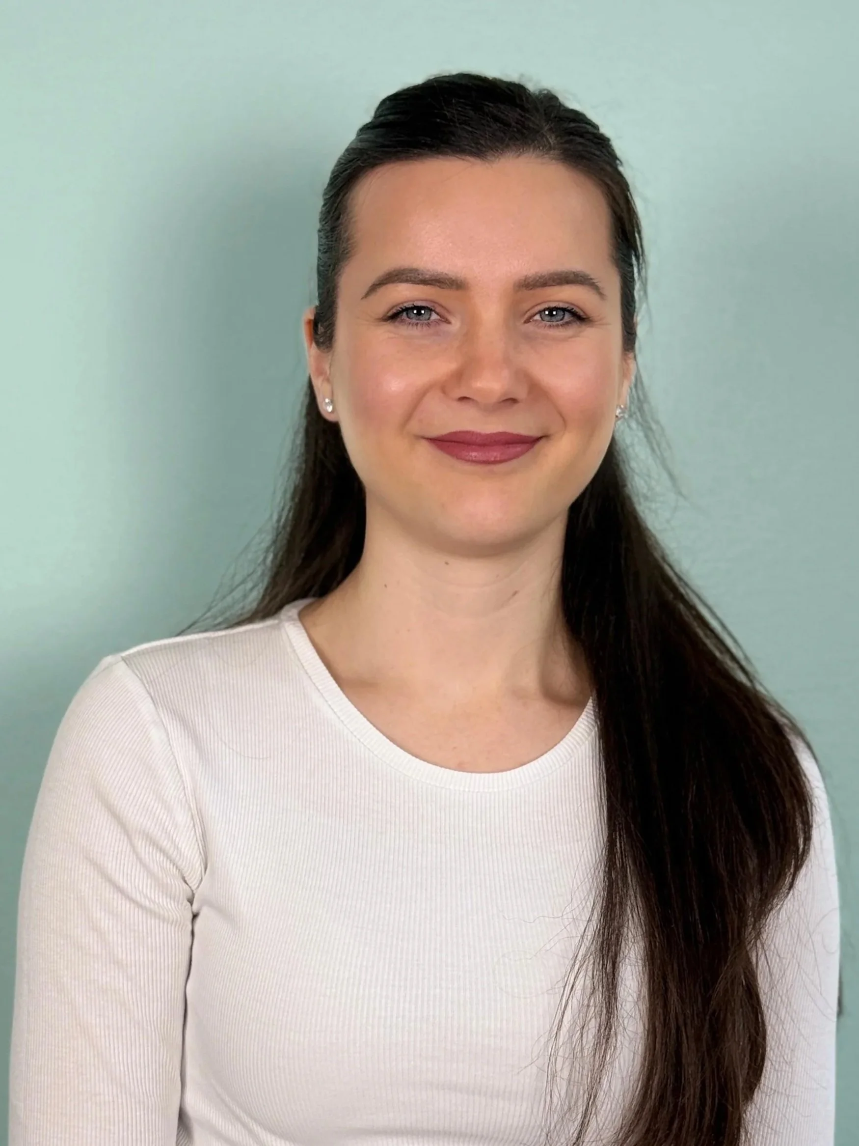 Junge Frau mit braunen Haaren, hellen Augen und einem weißen langärmligen Shirt, die vor einem blassen grünen Hintergrund lächelt.