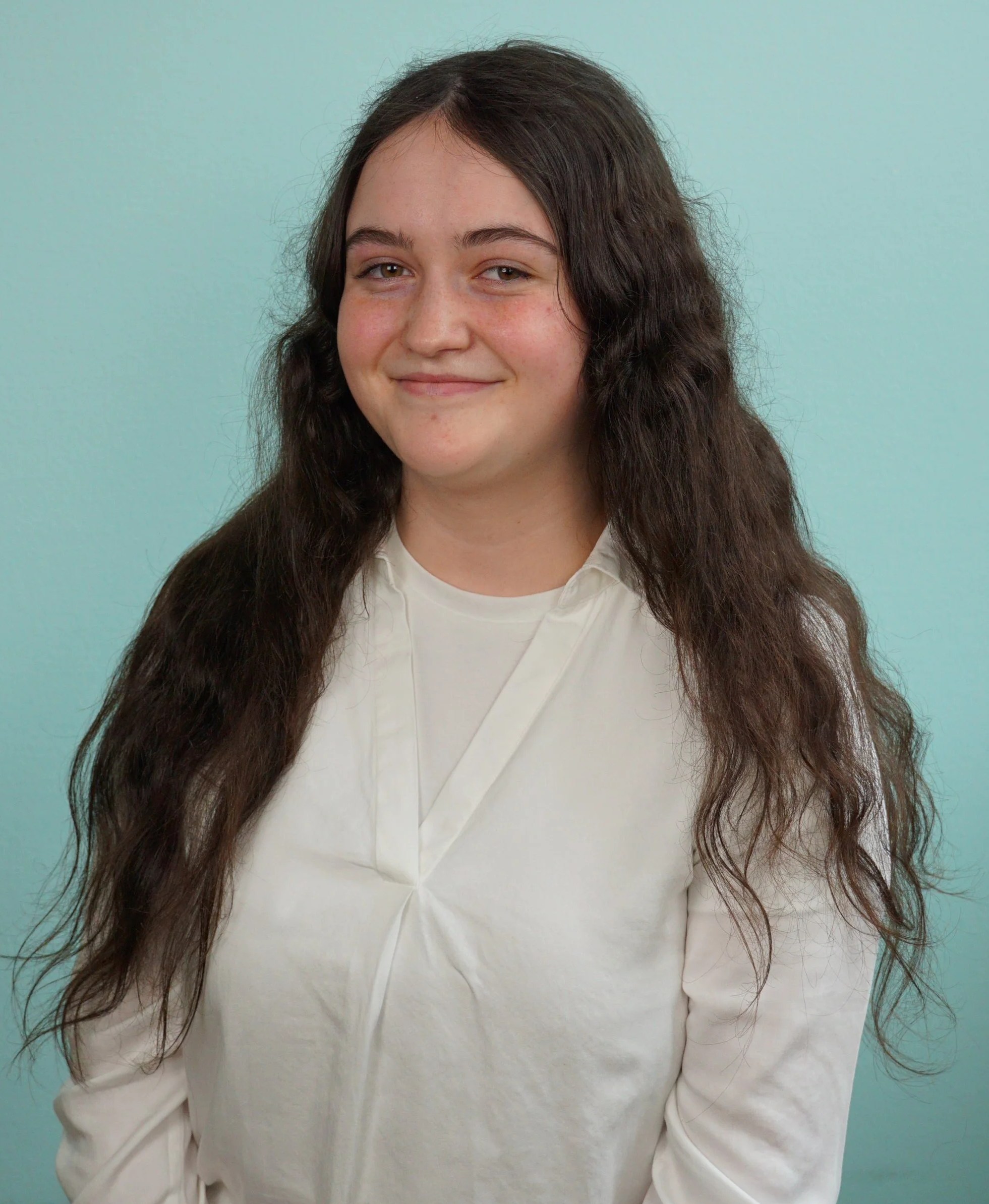 Junge Frau mit langem, dunklem, lockigem Haar, in weißer Kleidung, lächelnd vor hellem Himmelblau-Hintergrund.