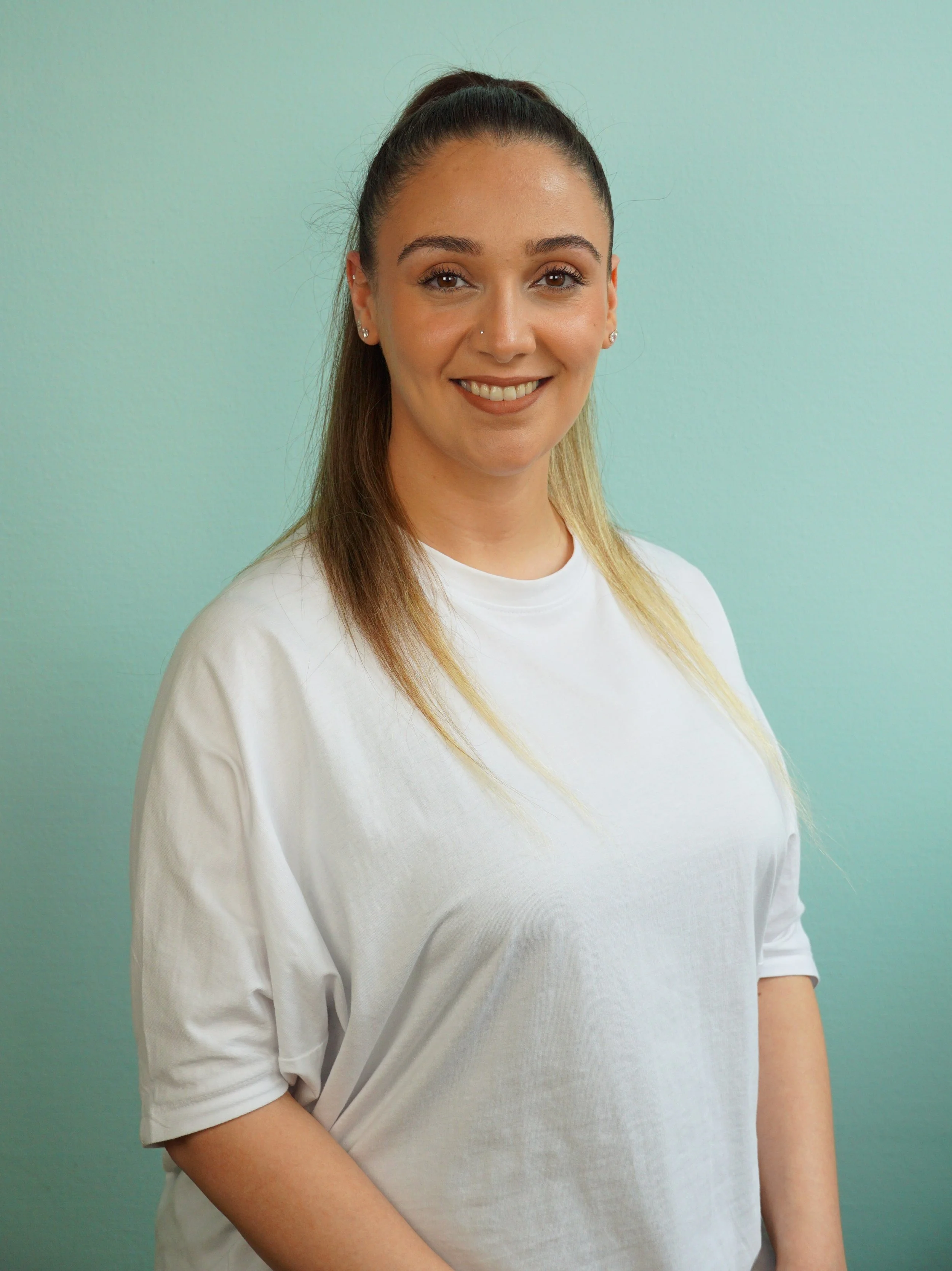 Ein Lächeln tragendes junges Frau mit dunklen, hochgesteckten Haaren, die einen weißen T-Shirt trägt, vor einem hellblauen Hintergrund.