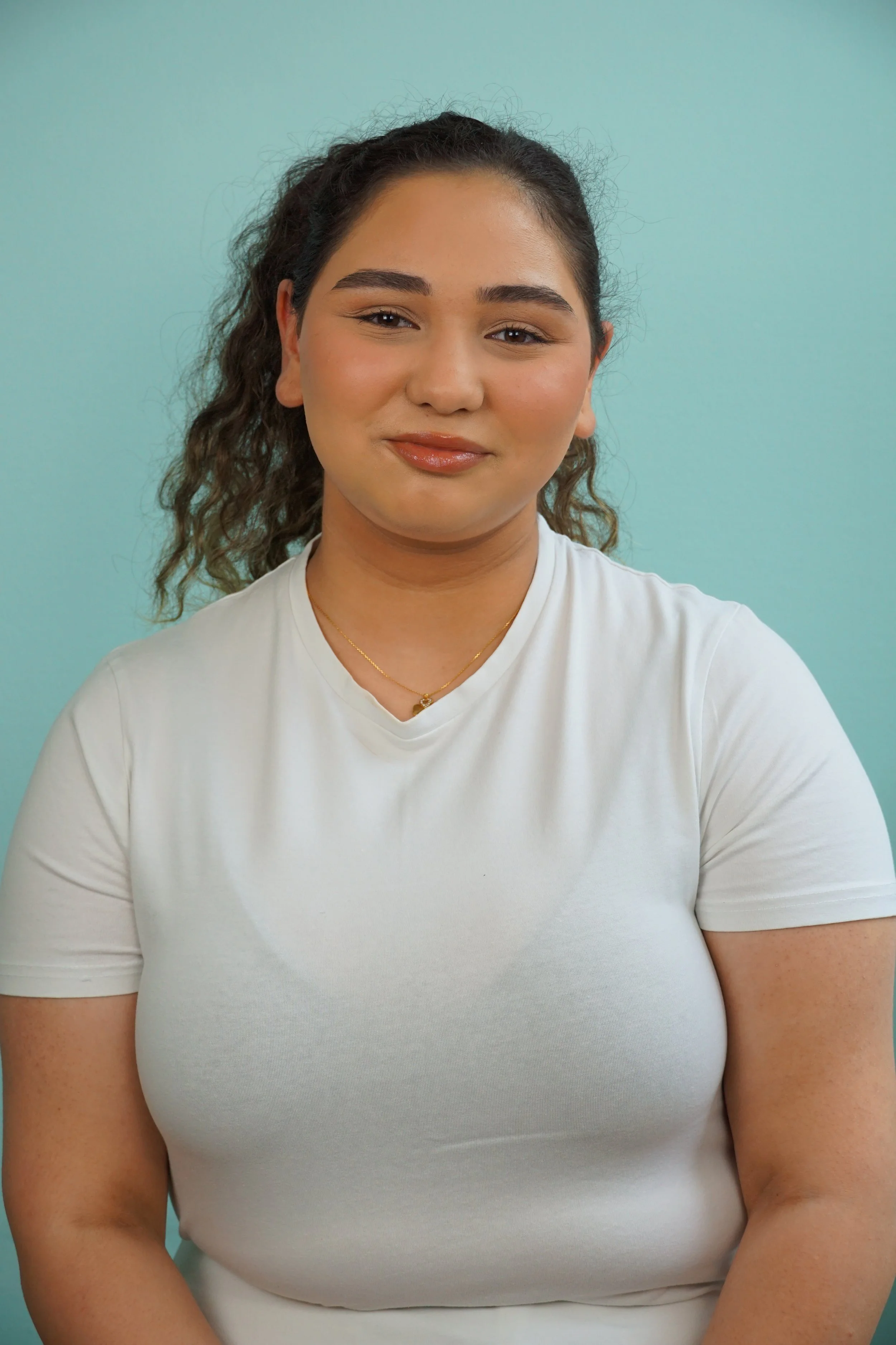 Porträt eines lächelnden jungen Mädchens mit dunklen, lockigen Haaren vor einem hellblauen Hintergrund, trägt ein weißes T-Shirt und eine goldene Halskette.