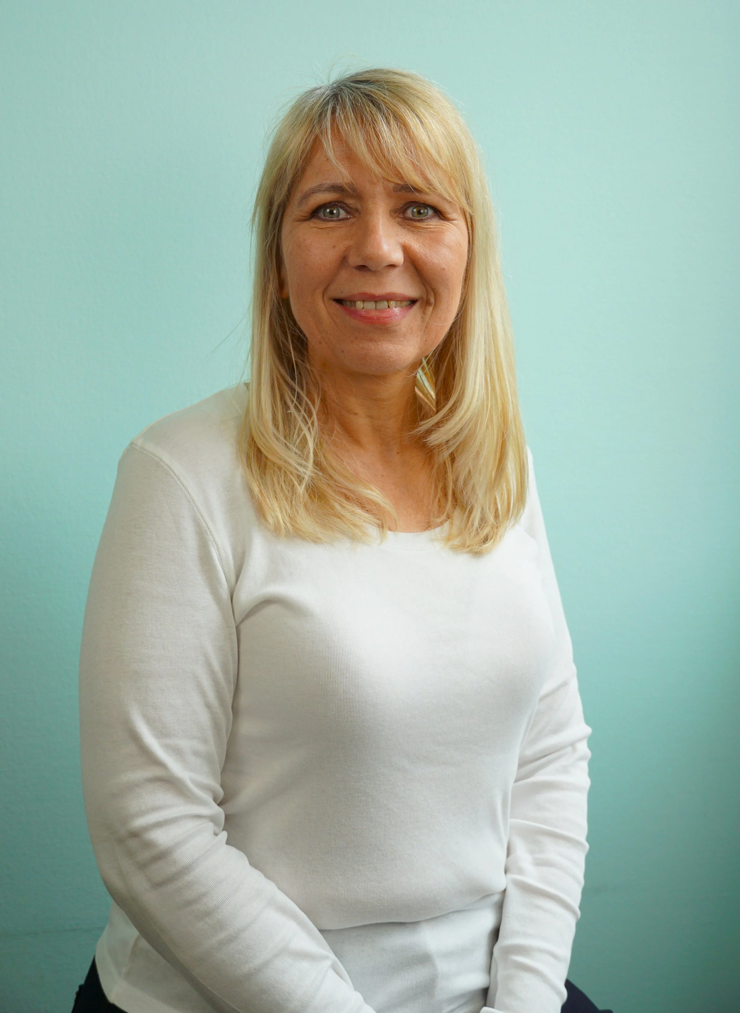 Ein lächelnder Mann mit blonden, schulterlangen Haaren trägt ein weißes Oberteil und sitzt vor einer hellblauen Wand.