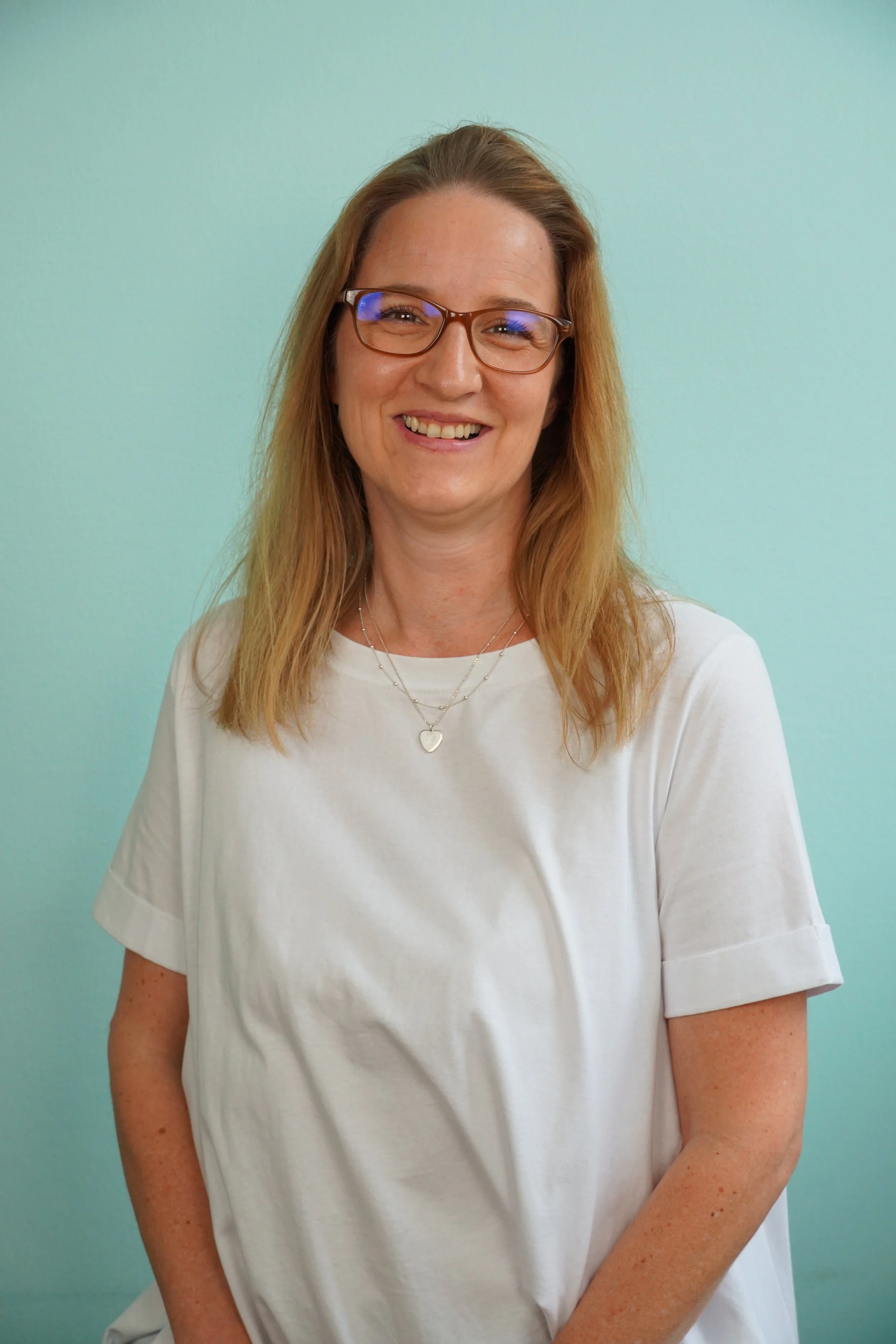 Ein lächelnder Mann mit roten Haaren, Brille und weißen T-Shirt vor einem hellblauen Hintergrund.