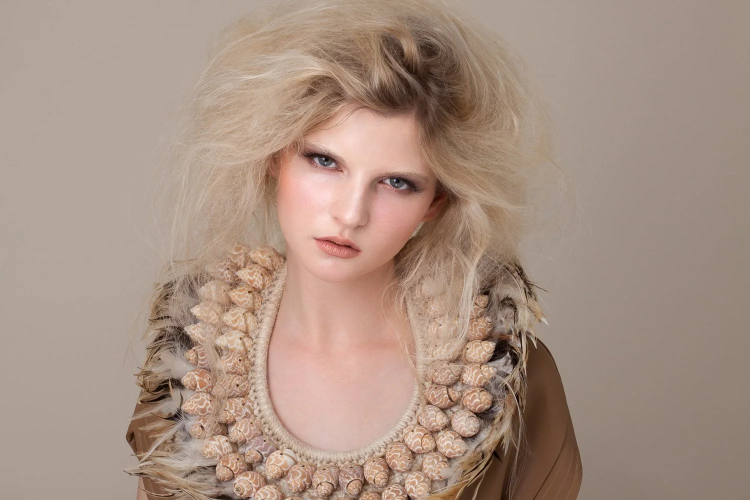 Close-up portrait of a model with tousled blonde hair and a statement shell and feather collar, styled and art directed by Laurence Trolet, fashion photographer